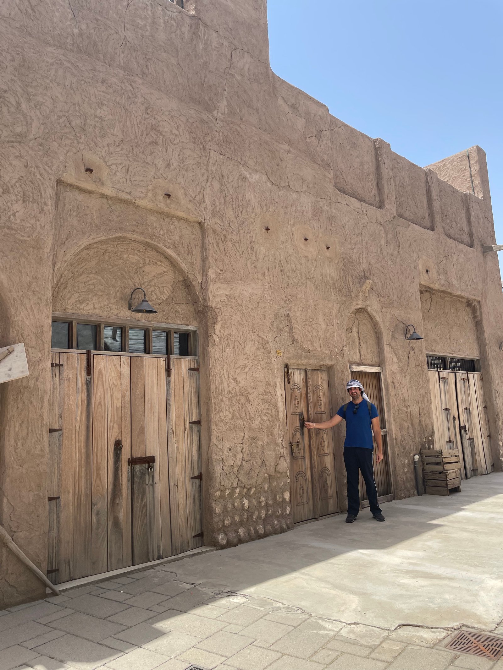 Homem em pé encostado na fachada de uma construção histórica de alvenaria em tons de terra com portas de madeira maciça e janelas tradicionais no distrito histórico de Al Seef, em Dubai.
