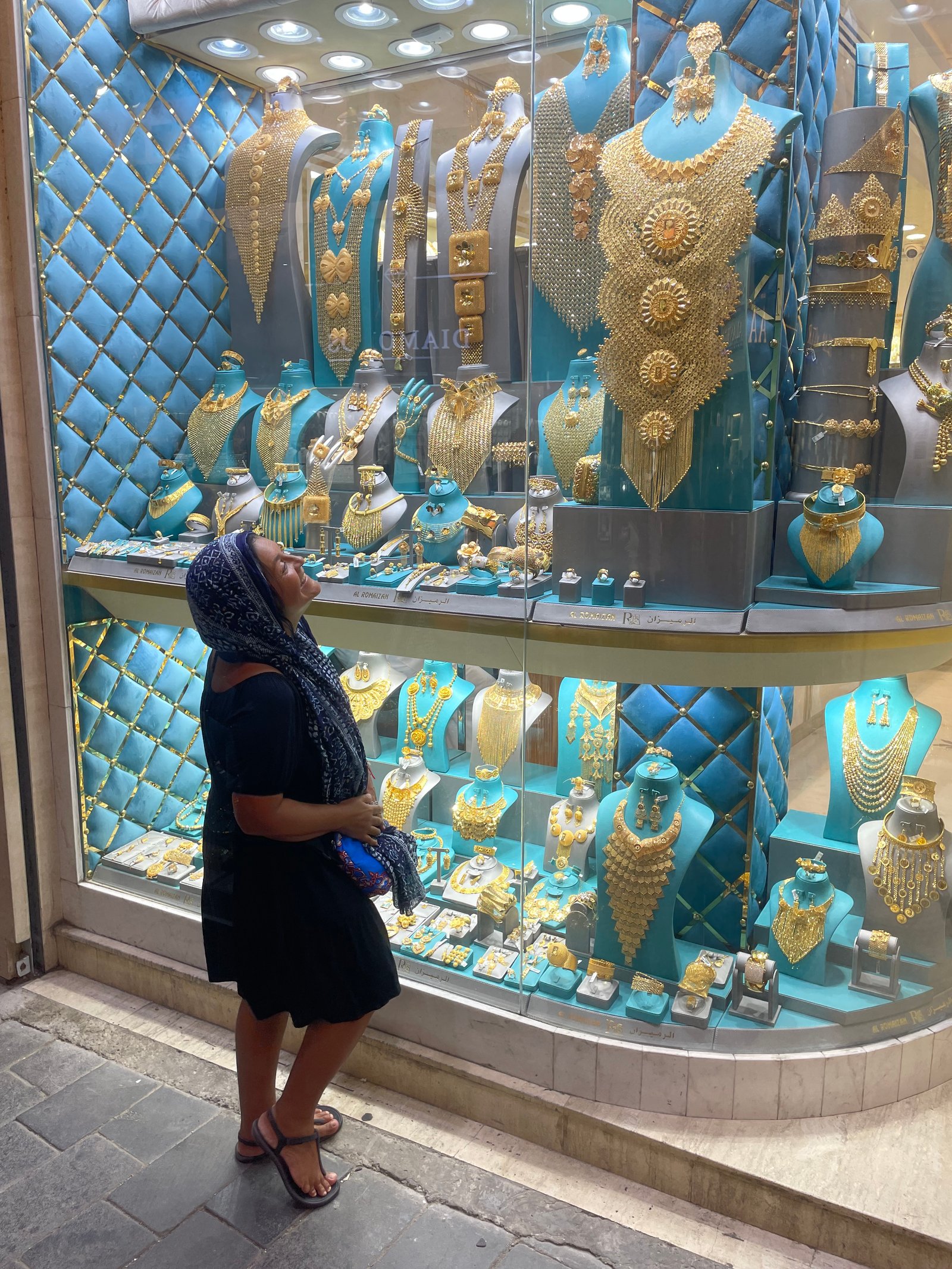 Mulher de vestido preto e lenço quadriculado preto e azul cobrindo a cabeça observando a vitrine ricamente iluminada de uma joalheria no tradicional Mercado de Ouro de Dubai (Gold Souk), exibindo dezenas de colares, brincos e adornos maciços de ouro amarelo contra um fundo de veludo azul capitonê