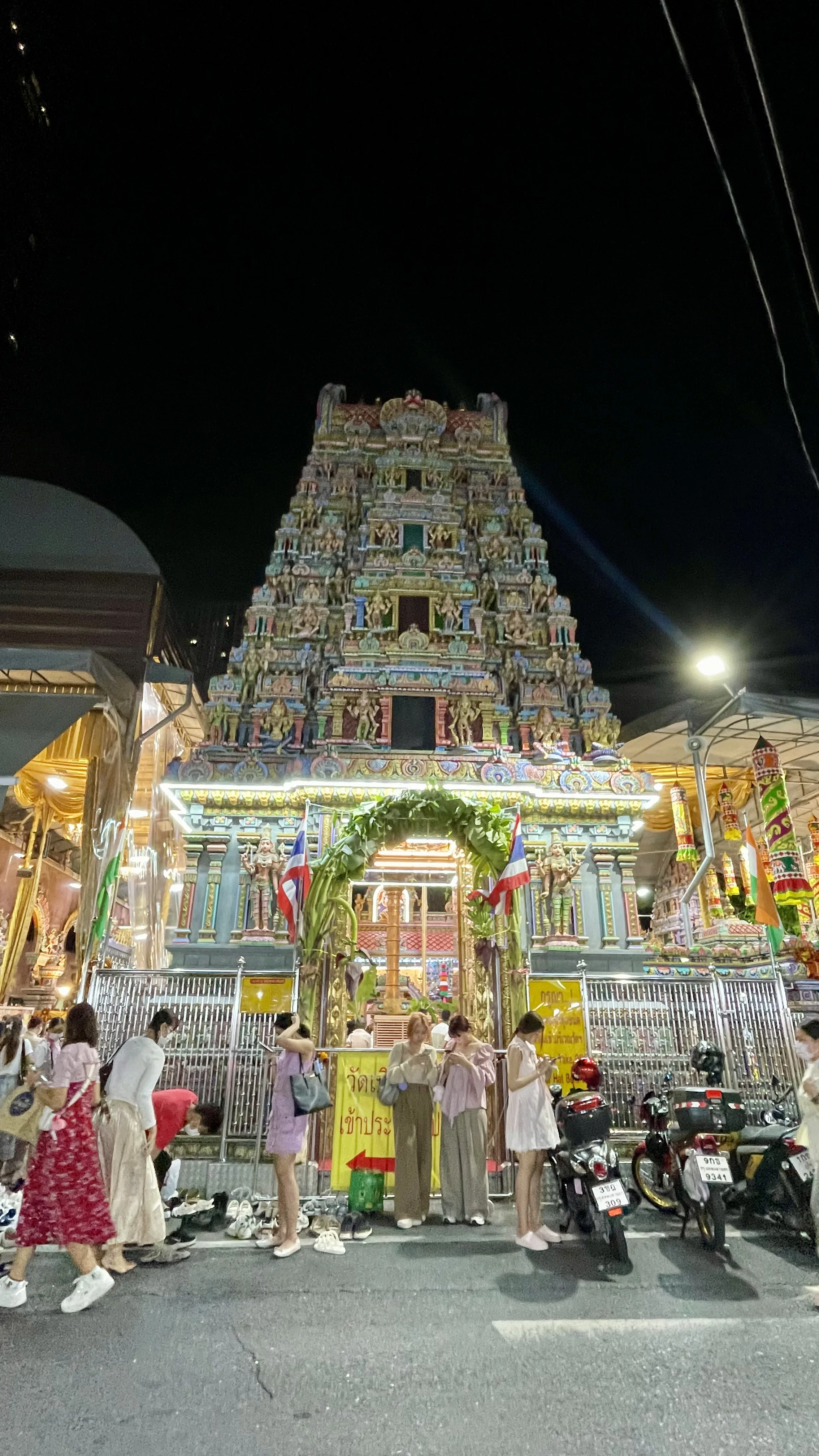 Fachada ricamente detalhada e iluminada à noite do Templo Hindu Sri Mariamman em Banguecoque, com pessoas em frente