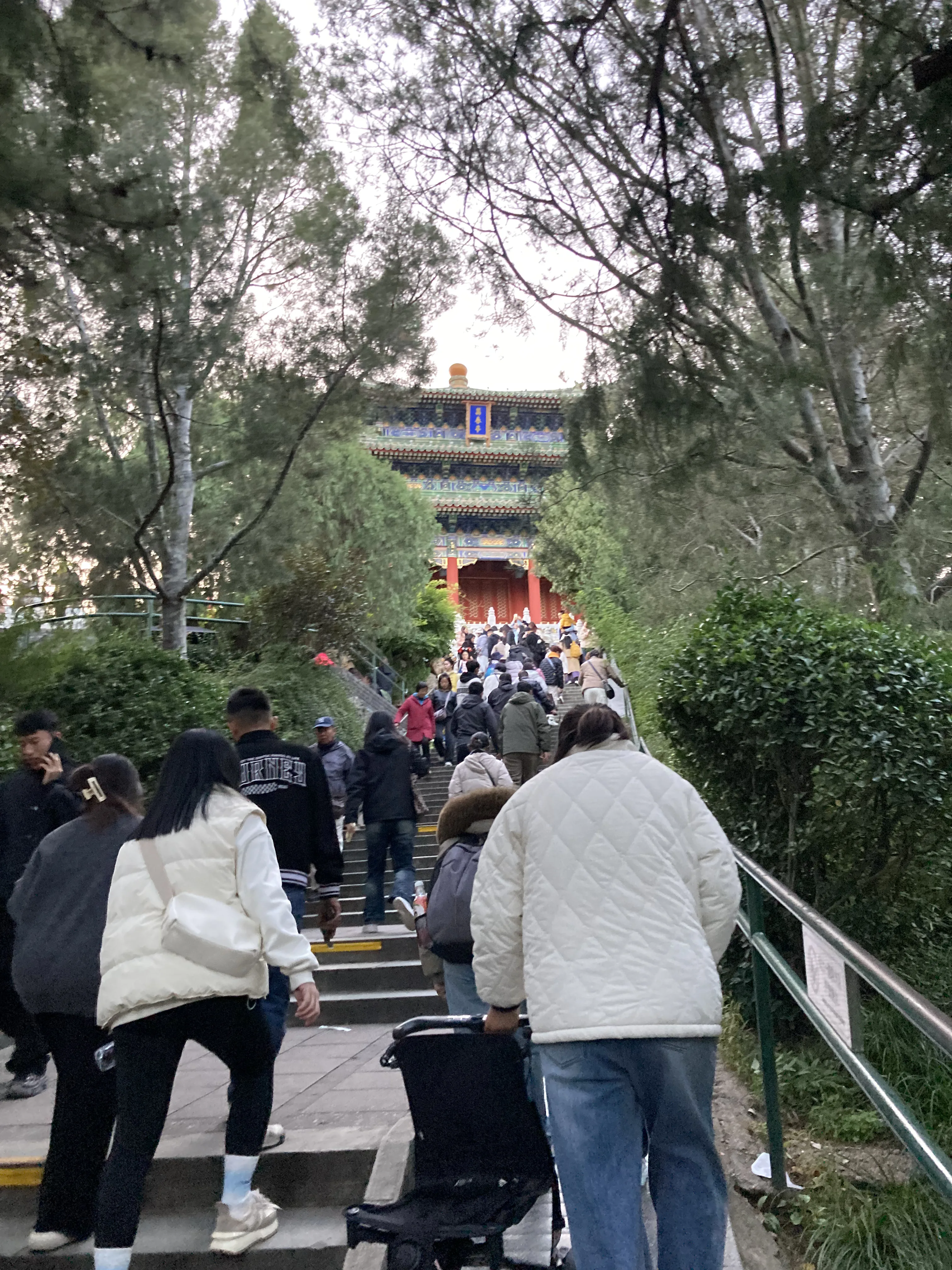Pessoas sobem escadaria em parque arborizado em Pequim em direção a pavilhão tradicional ao fundo.
