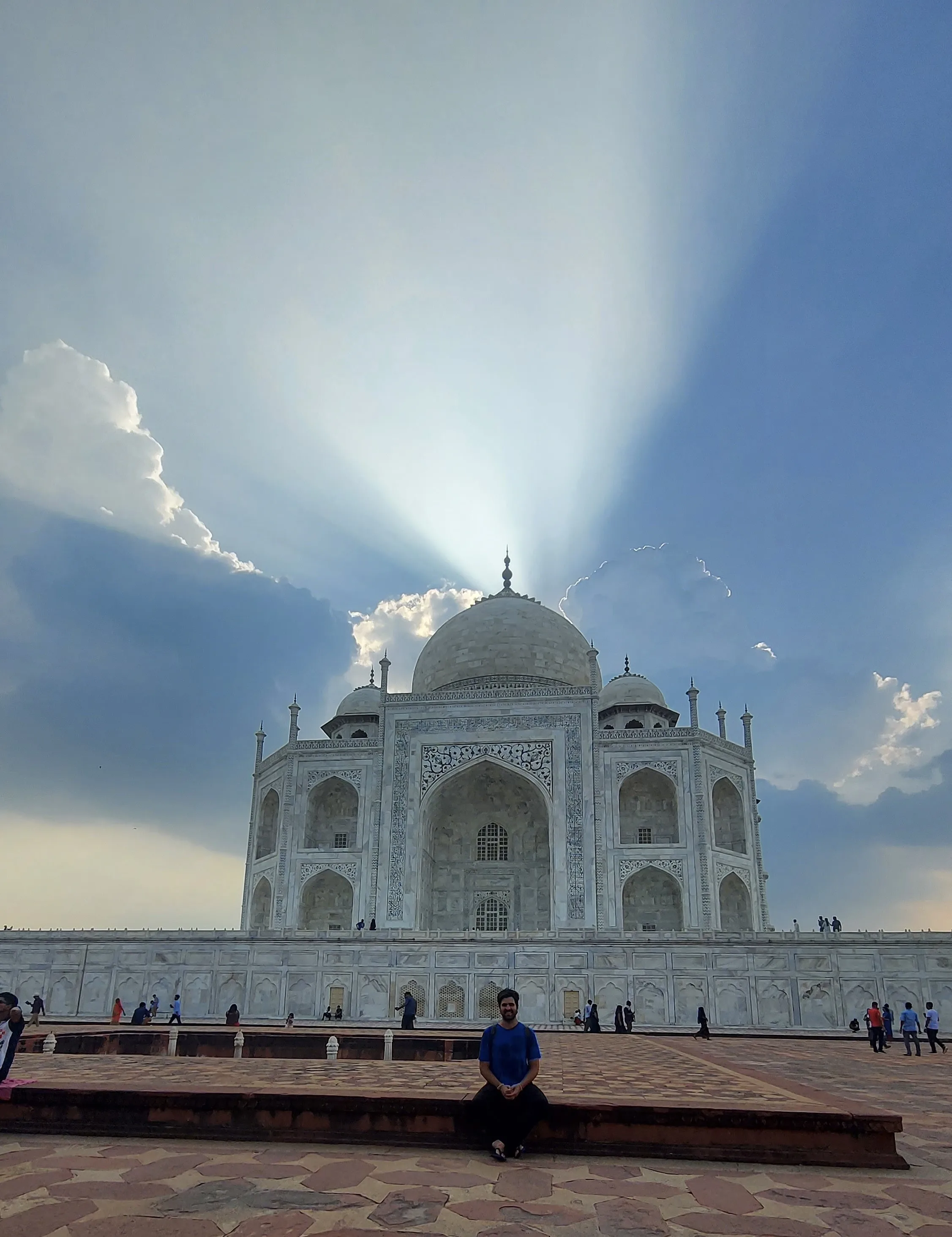 Pessoa sentada na mureta do pátio diante do Taj Mahal, com a monumental cúpula de mármore branco em destaque sob um céu parcialmente nublado e raios de luz solar que se projetam para cima.