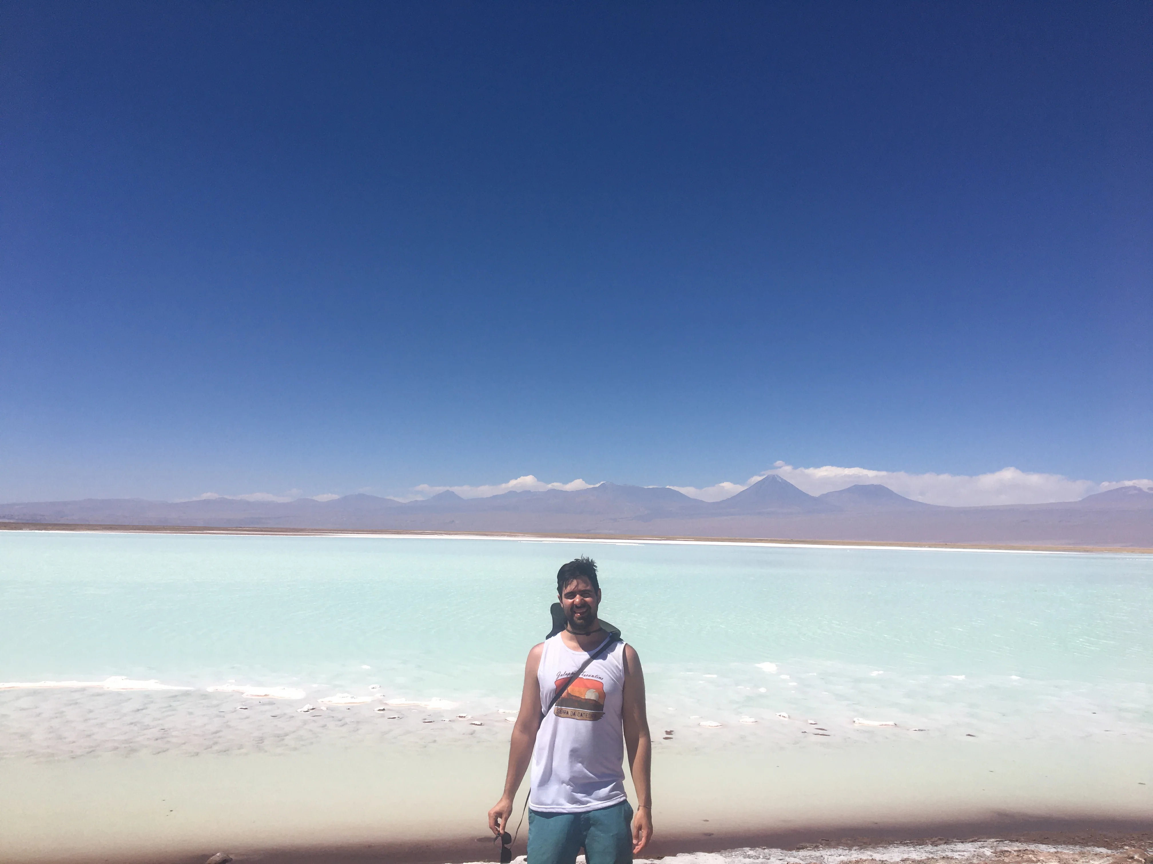 Homem sorri em pé na borda de sal branco diante das águas turquesas e montanhas ao fundo na Laguna Tebenquiche no Chile.