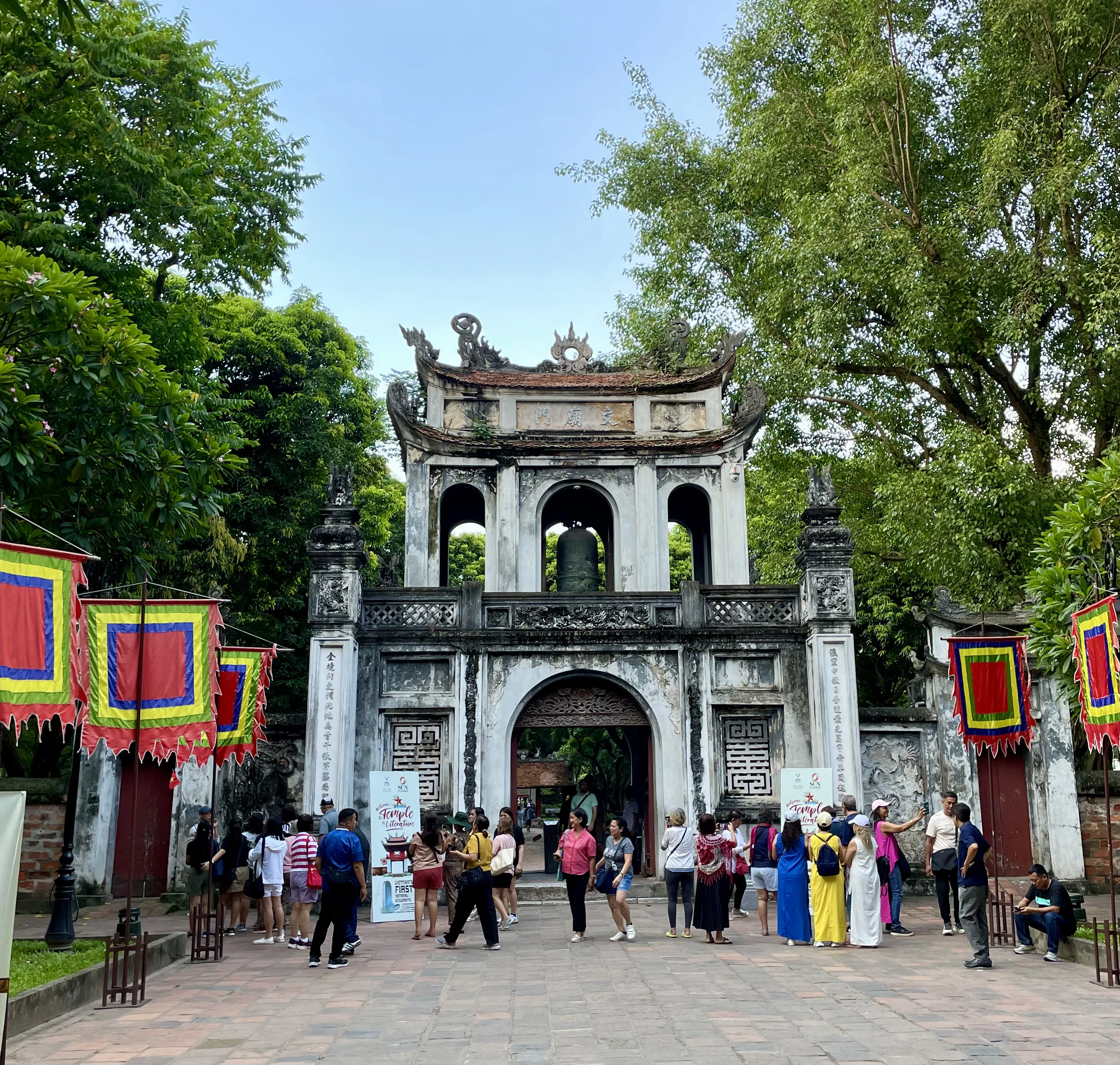 Vista frontal do portão histórico de entrada do Templo da Literatura em Hanói, uma estrutura de alvenaria cinzenta envelhecida com telhado tradicional recurvado onde se avista um sino de bronze pendurado no arco superior. A praça pavimentada em frente está repleta de turistas caminhando e conversando, ladeada por árvores frondosas e bandeiras vietnamitas coloridas penduradas em hastes.