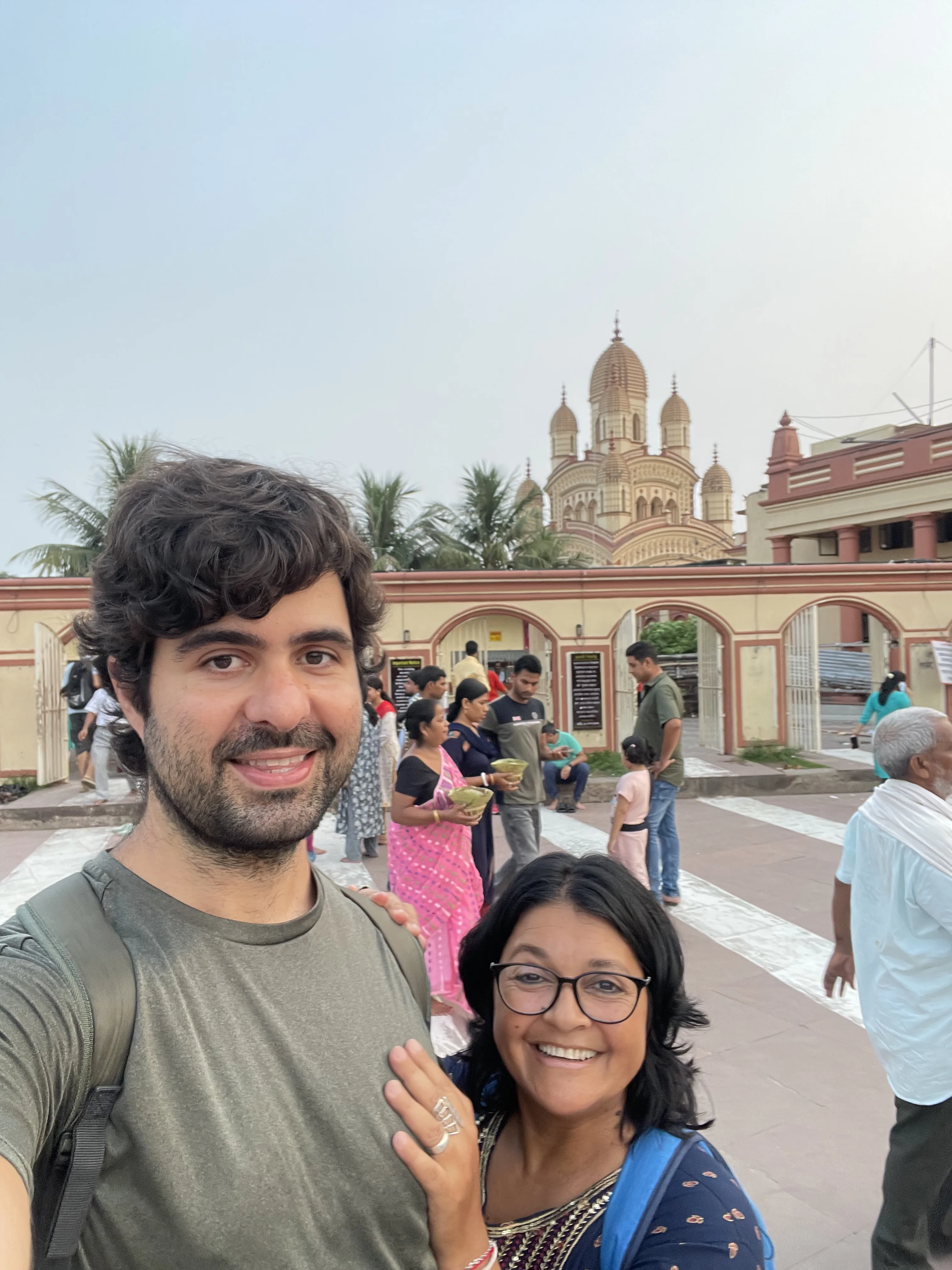 Um homem e uma mulher sorridentes em primeiro plano fazendo selfie no pátio externo do Templo Dakshineswar Kali, com visitantes caminhando e as cúpulas ornamentadas do templo indiano ao fundo
