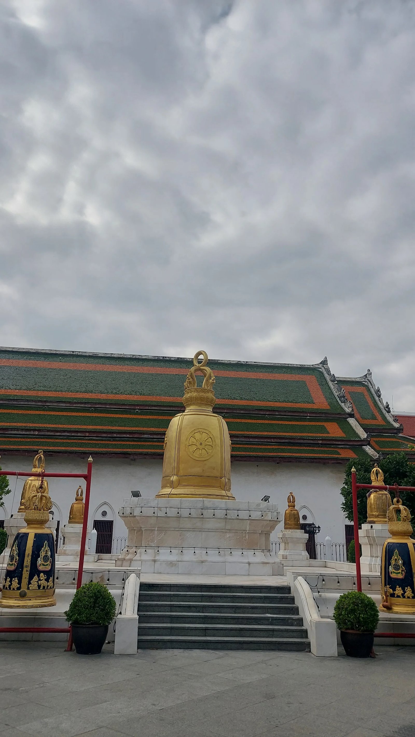 Grande estrutura de sino dourado e sinos menores decorados no complexo do templo em Banguecoque, sob um céu nublado.