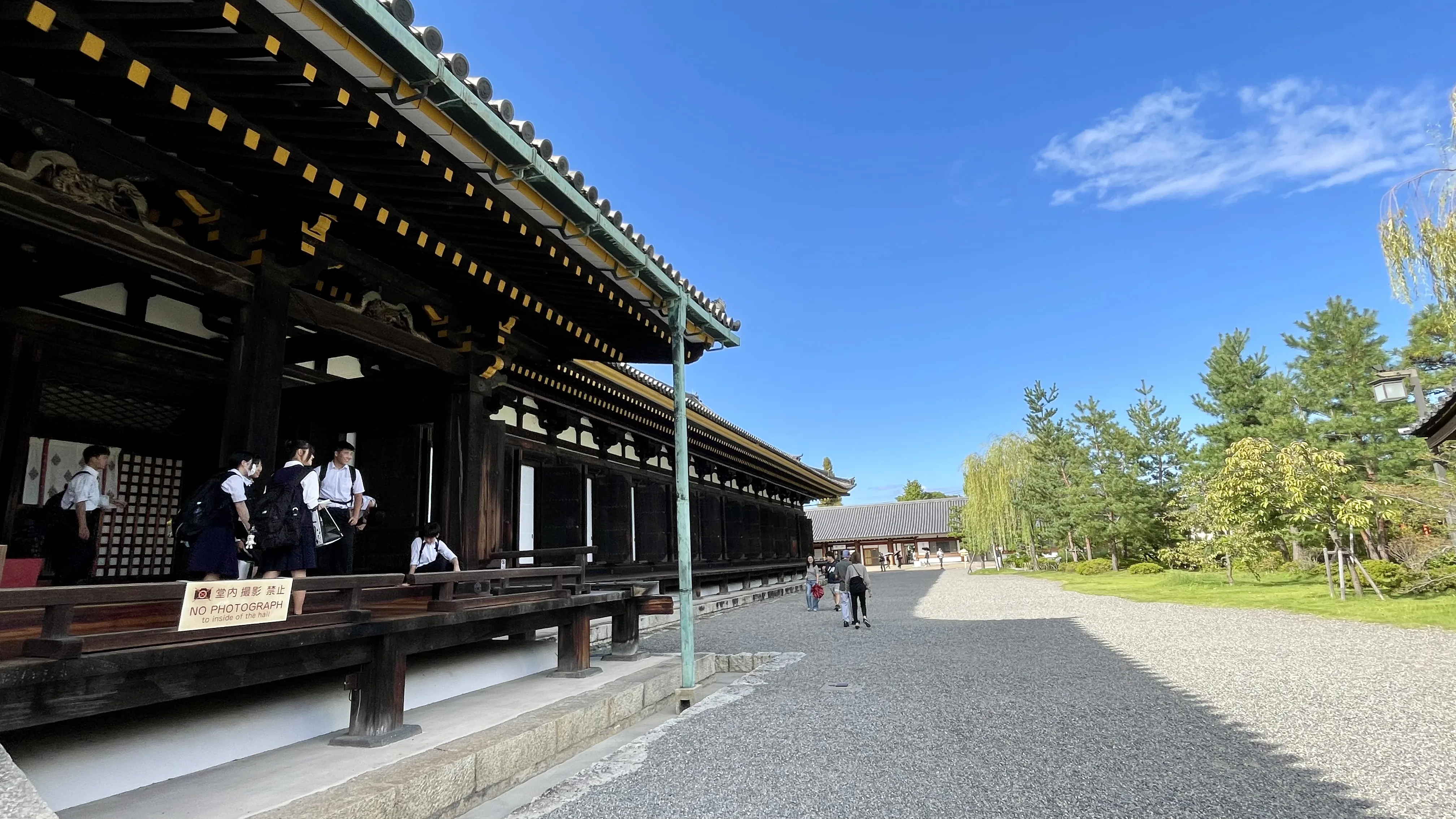 Estudantes e visitantes no pátio e na passarela externa do Salão Sanjusangen-do, templo das 1001 estátuas de Kannon em Kyoto, sob céu azul.