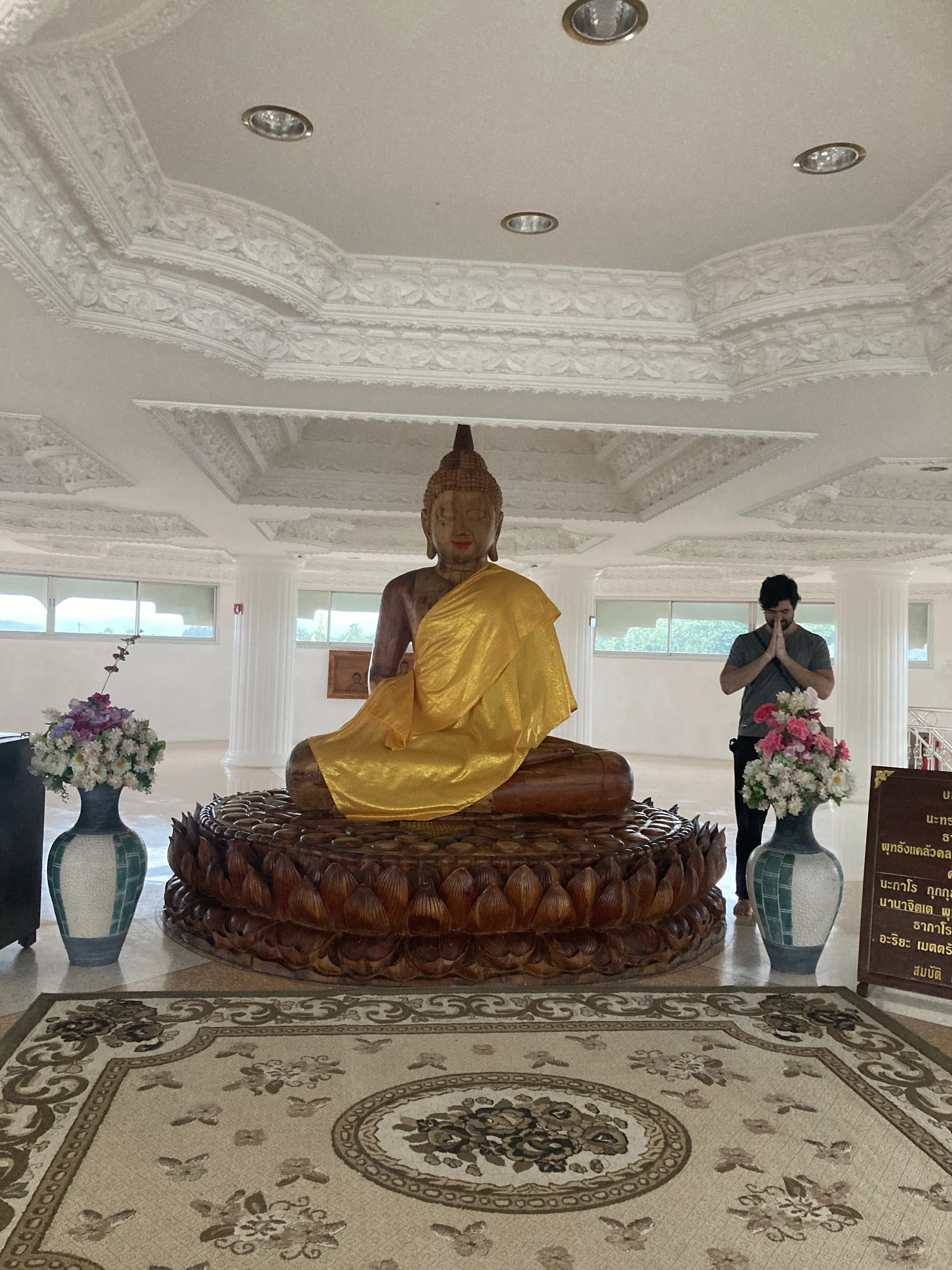 Estátua de Buda de madeira adornada com túnica amarela e cercada por cobras nagas, posicionada entre vasos de flores em um templo.