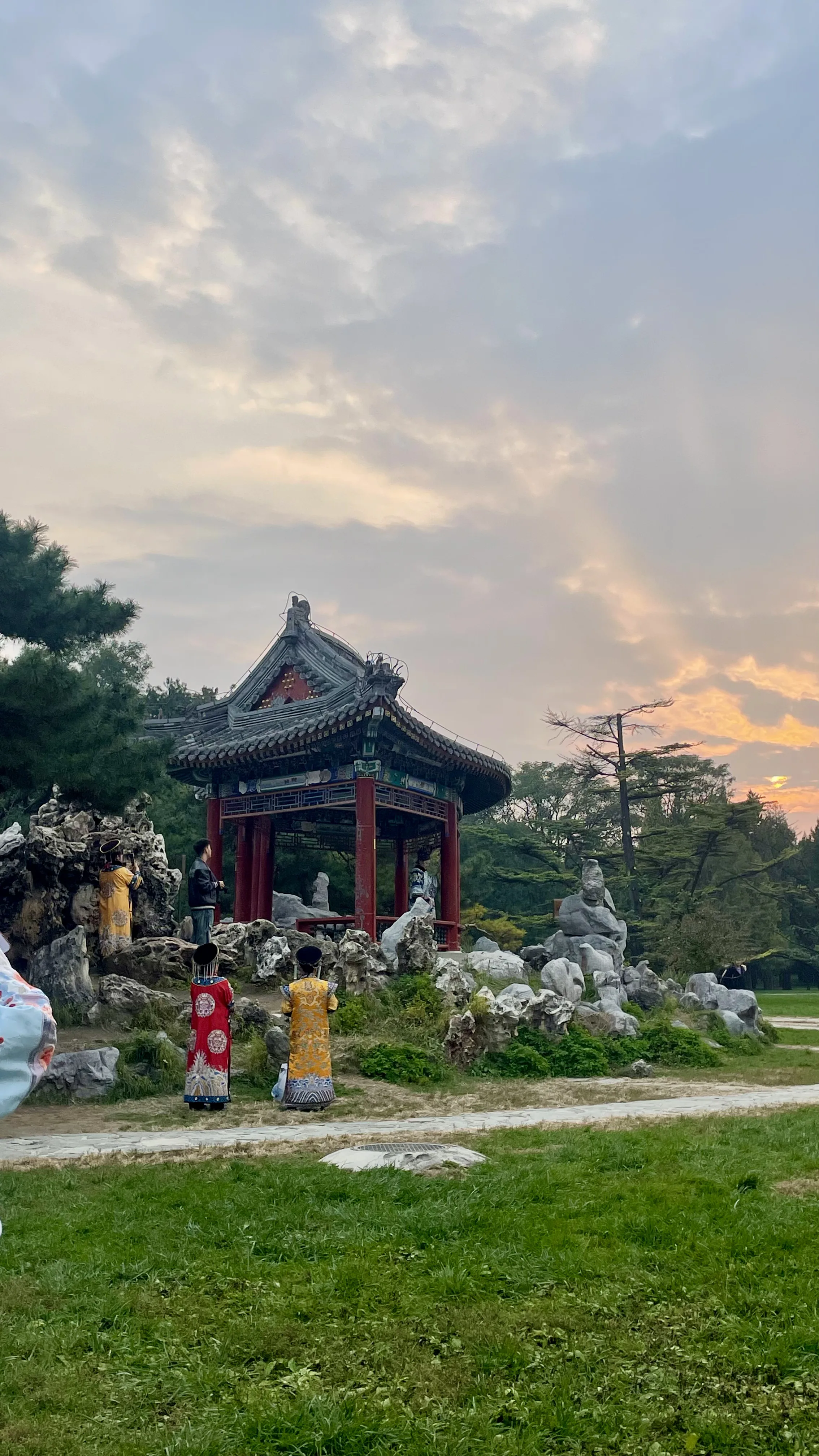 Pessoas vestidas com trajes tradicionais chineses caminham perto de pavilhão e formações rochosas ao entardecer no Templo do Céu em Pequim.