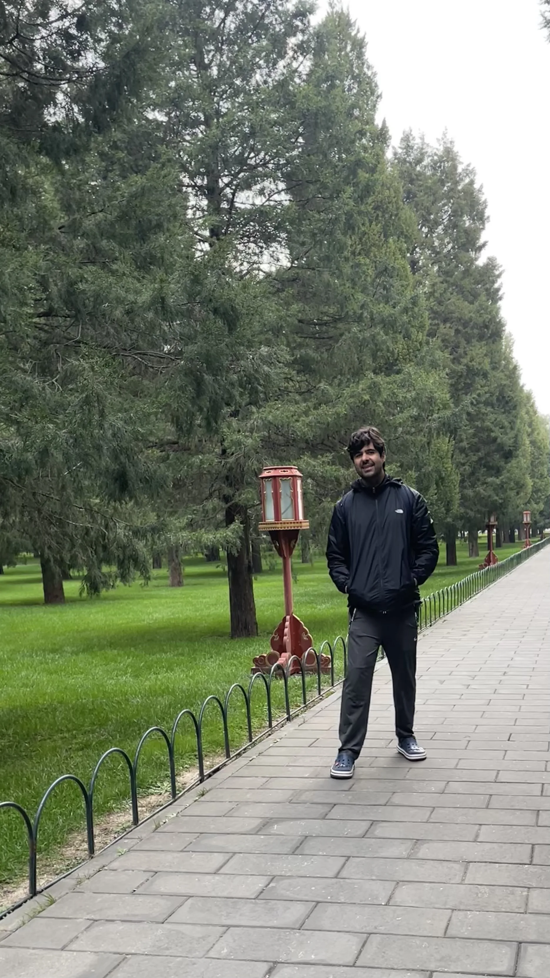 Homem sorri em pé em calçada pavimentada ladeada por gramado verde e árvores altas no Templo do Céu em Pequim.