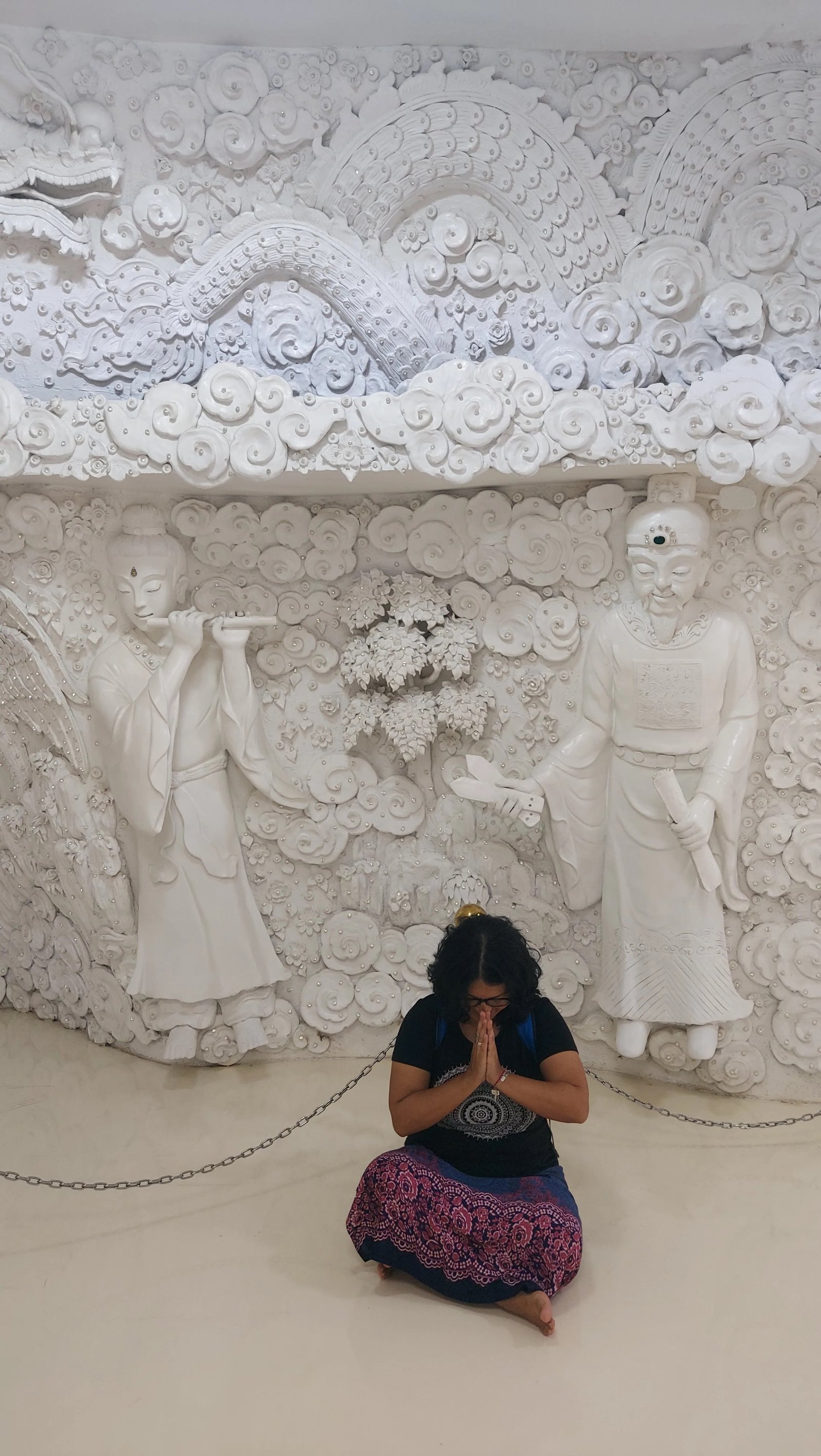 Mulher sentada com as mãos juntas em gesto de oração diante de relevos esculpidos em branco nas paredes internas do templo em Chiang Rai.
