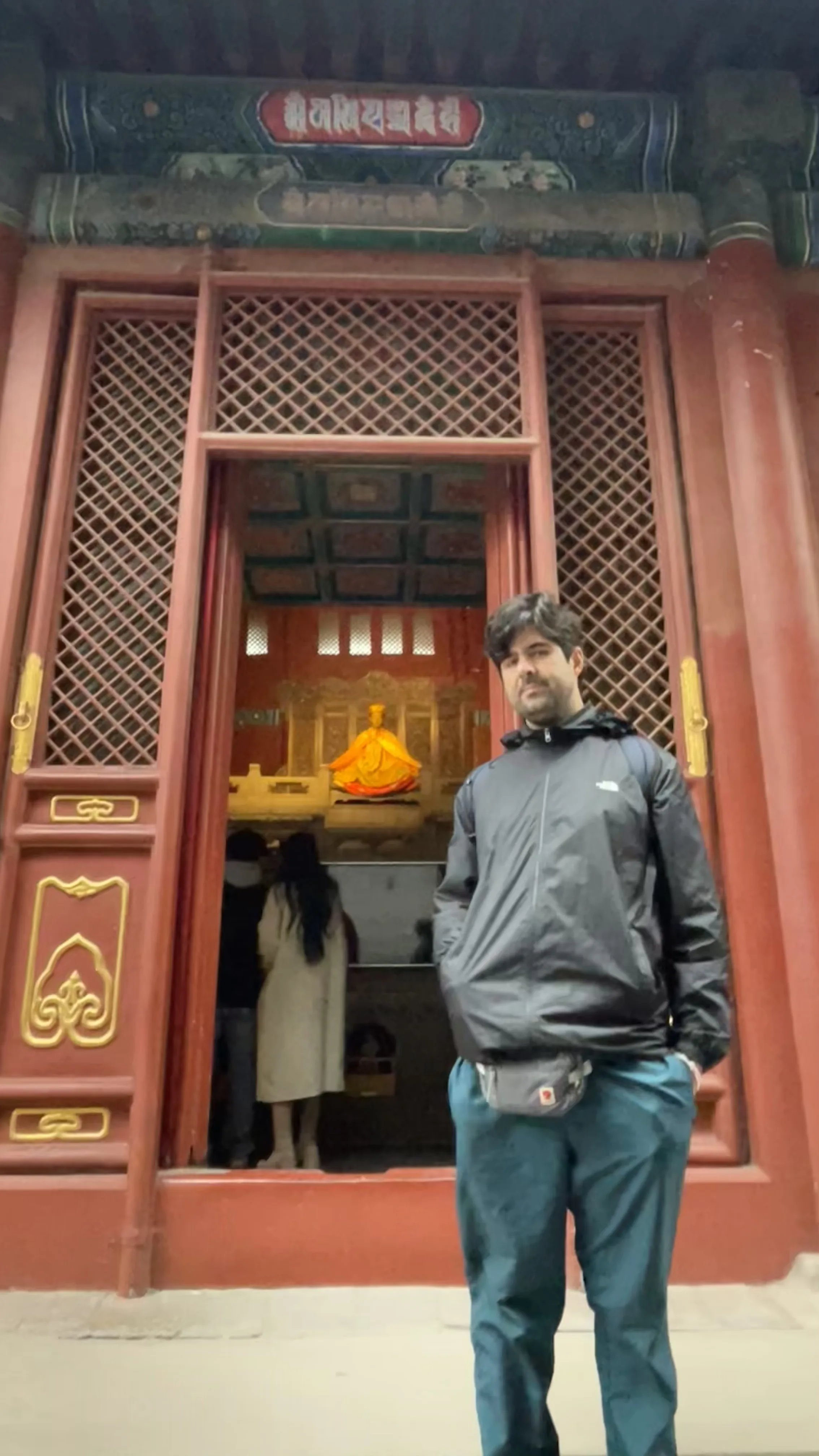 Homem em pé na entrada de templo com portas de madeira vermelha e treliças exibindo estátua dourada ao fundo em Pequim.