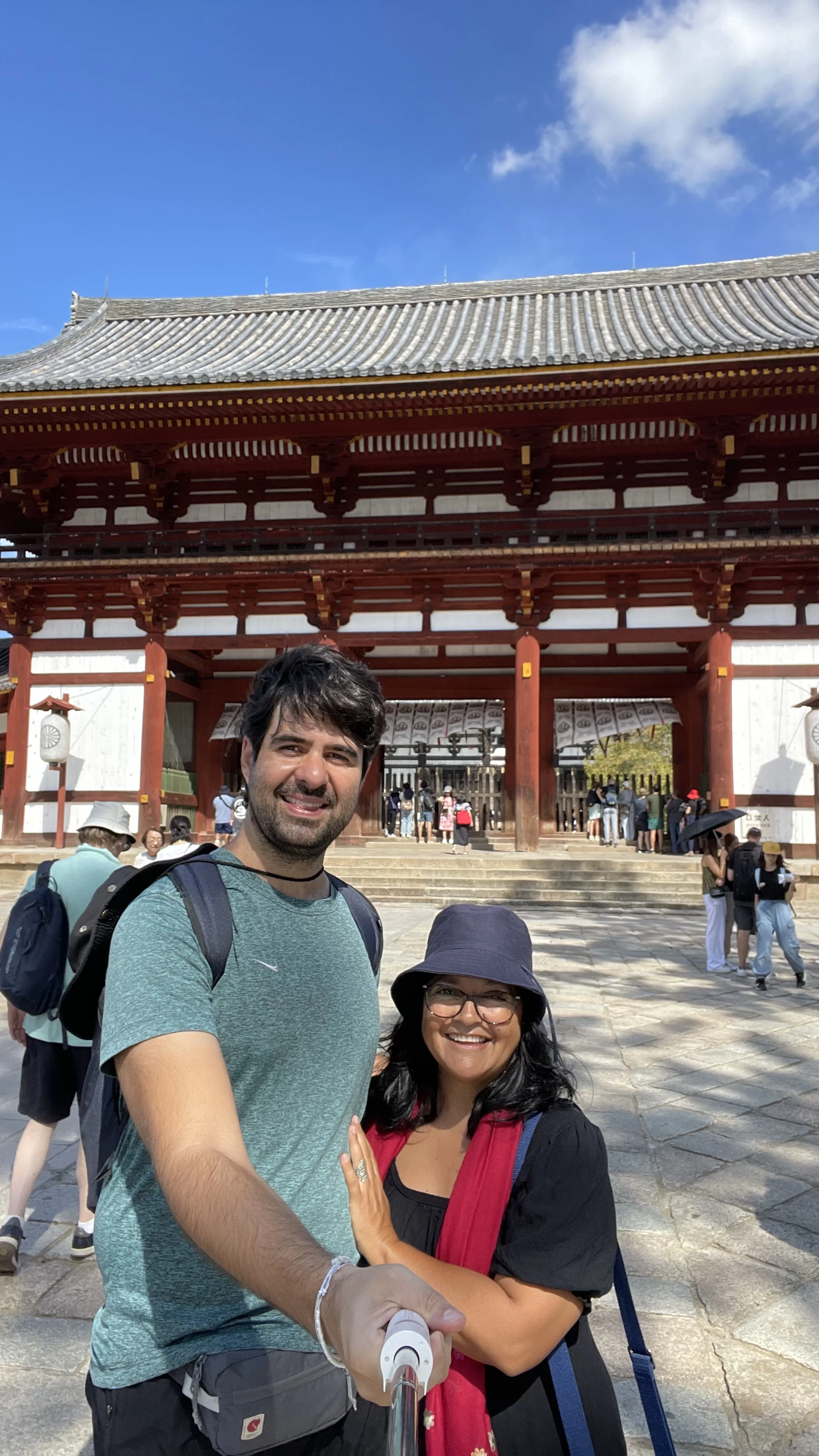 Turistas posam para foto no pátio de pedra do portão Chūmon no templo Tōdai-ji, em Nara. A estrutura tradicional apresenta colunas de madeira vermelha, paredes brancas e um telhado cinza ornamentado sob um céu azul