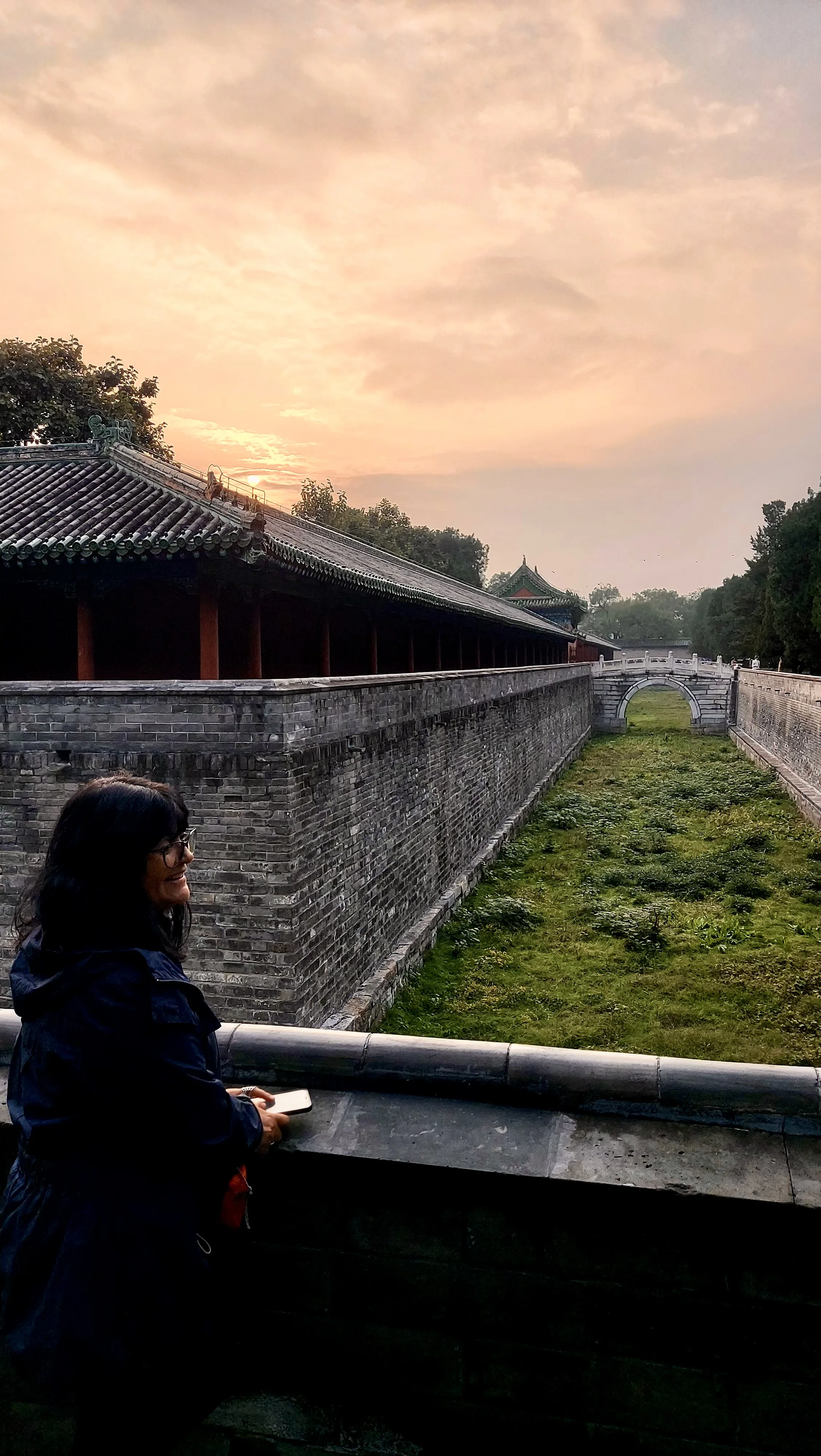 Mulher de perfil apoia-se em parapeito com vista para muralha antiga, fosso verde e ponte em arco sob o pôr do sol em Pequim.