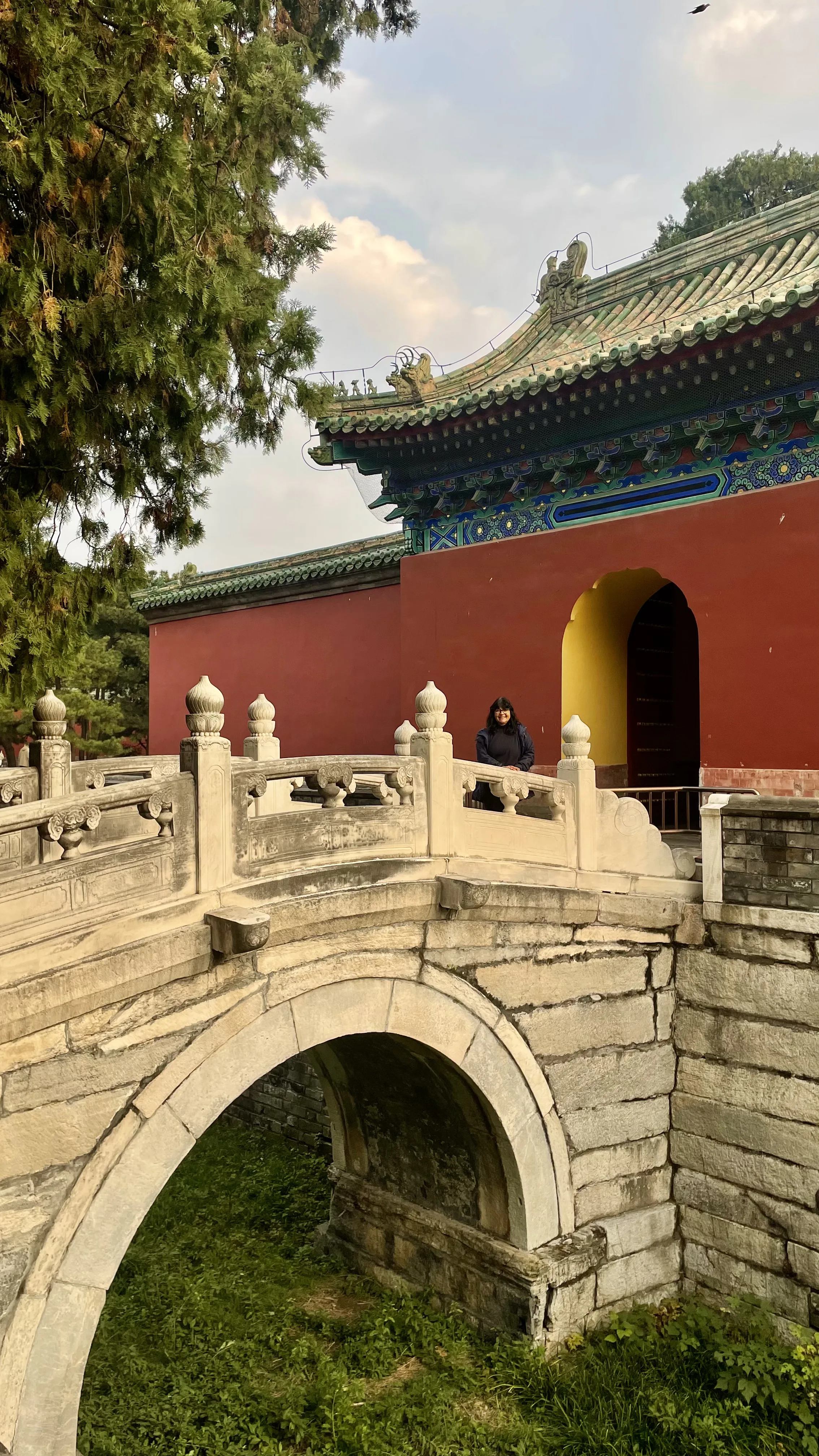 Mulher sorri em pé junto a ponte de pedra em arco com balaustrada ornamentada em frente a edifício histórico de paredes vermelhas e telhado verde em Pequim.