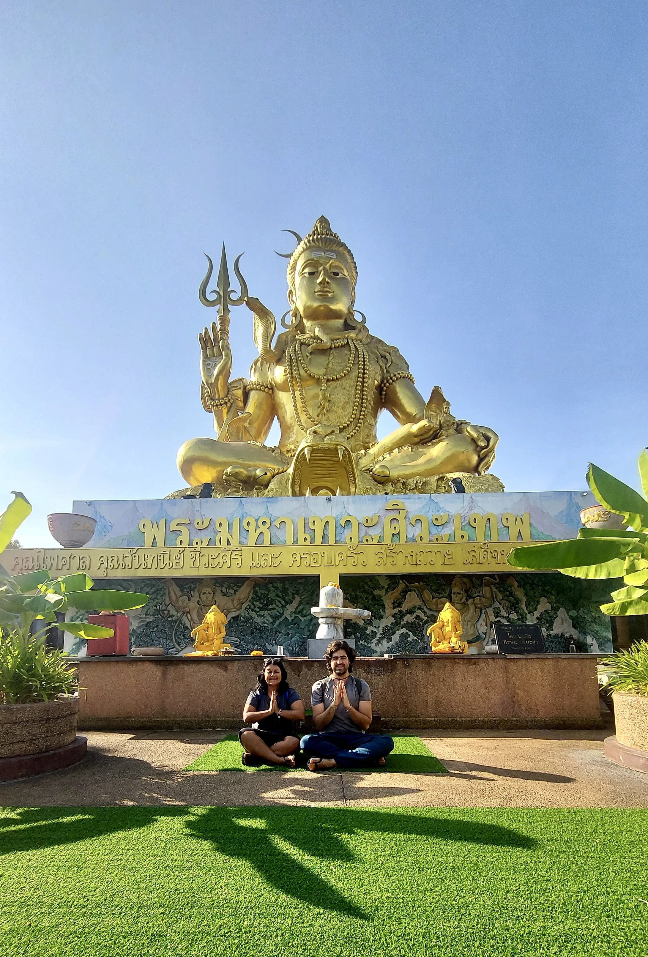 Casal sentado com as mãos unidas em reverência diante da imponente estátua dourada de Shiva no templo Wihan Sadet Pho, na Tailândia