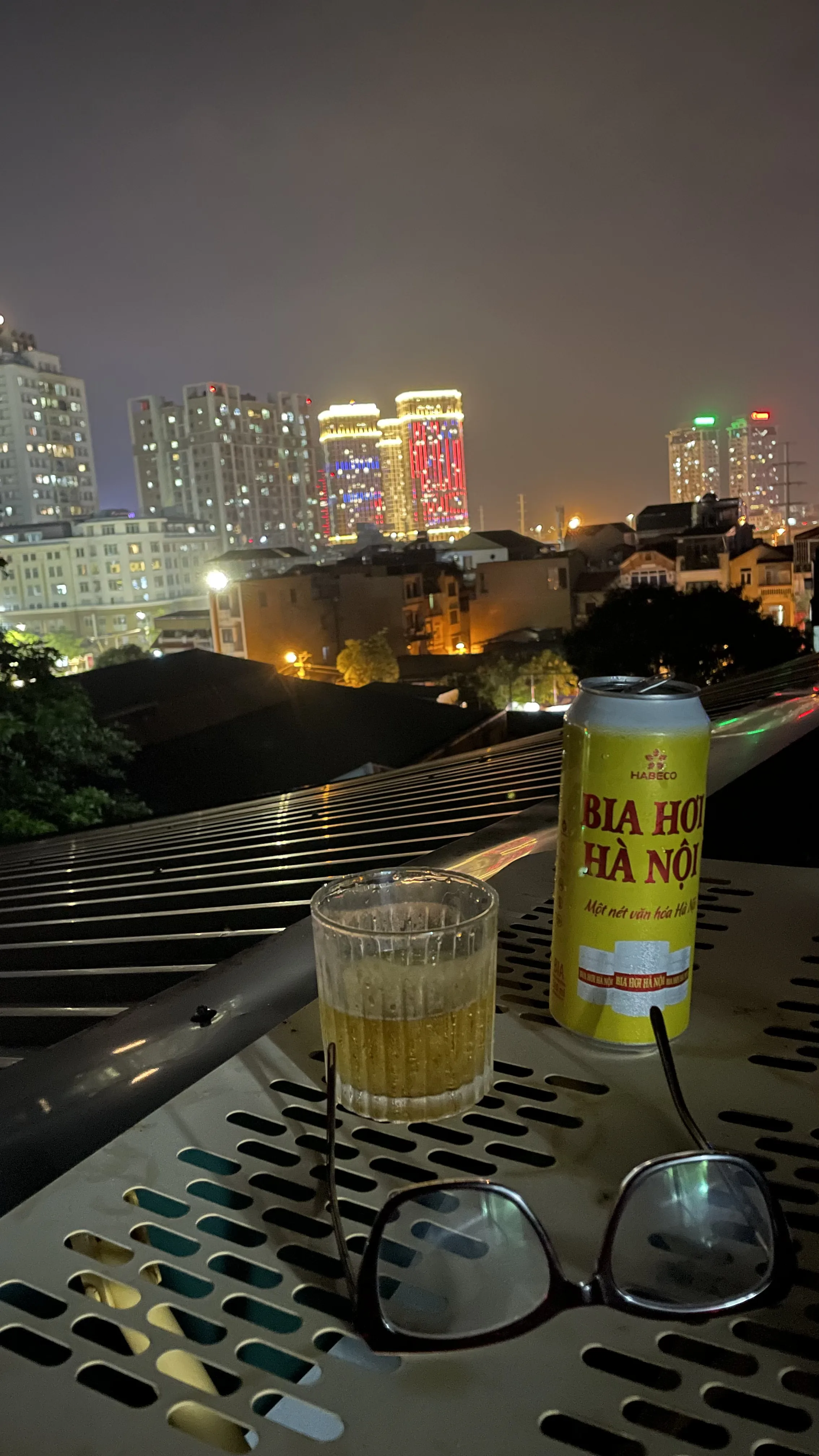 Cena noturna vista de um terraço em Hanói, com uma lata amarela de cerveja Bia Hà Nội, um copo de vidro com bebida alcoólica e óculos de grau apoiados sobre uma superfície metálica perfurada, tendo ao fundo a linha do horizonte da cidade iluminada por prédios altos.