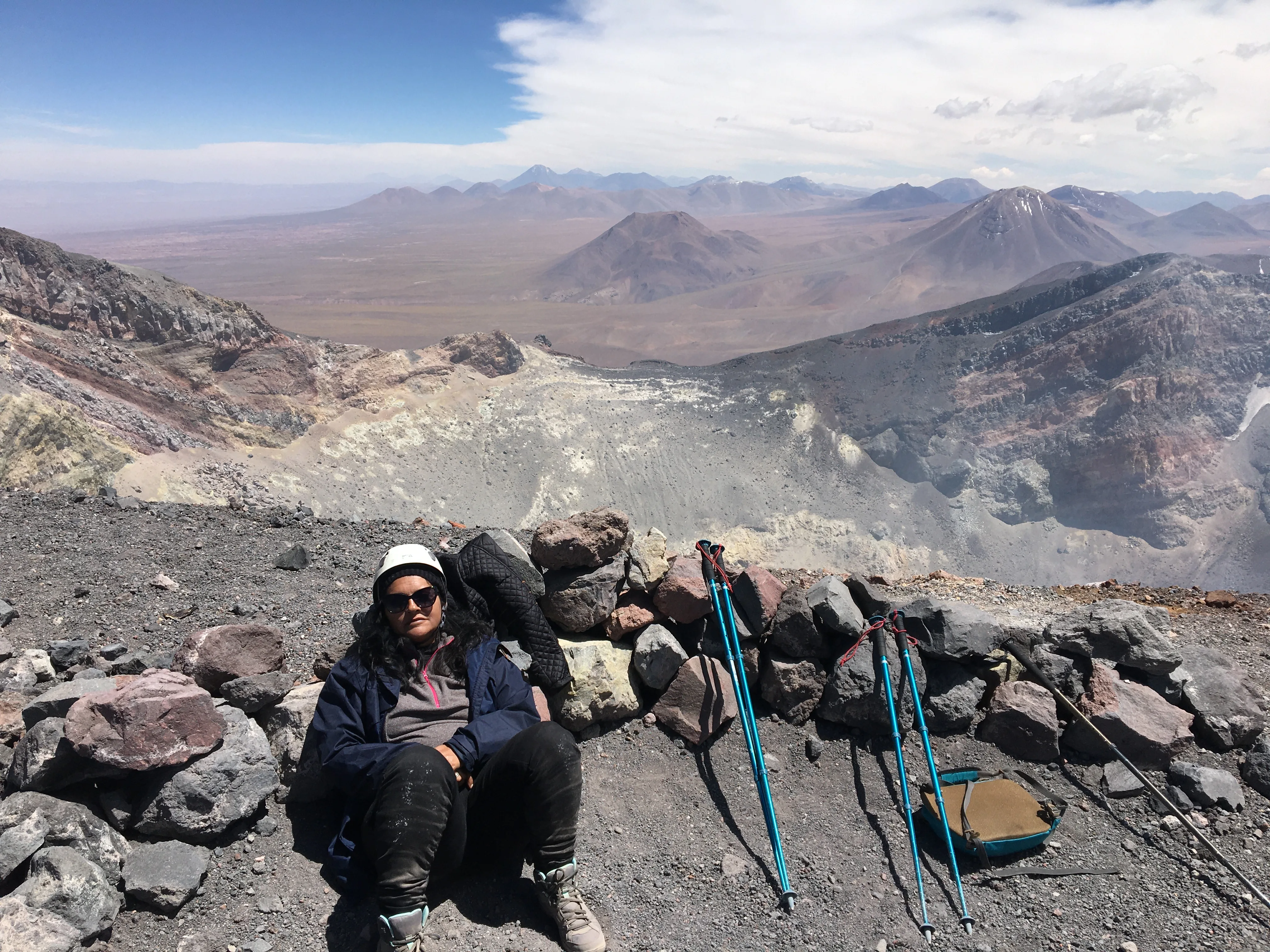 Mulher sentada entre pedras com equipamentos e bastões de trilha no cume do Vulcão Lascar, no Chile.