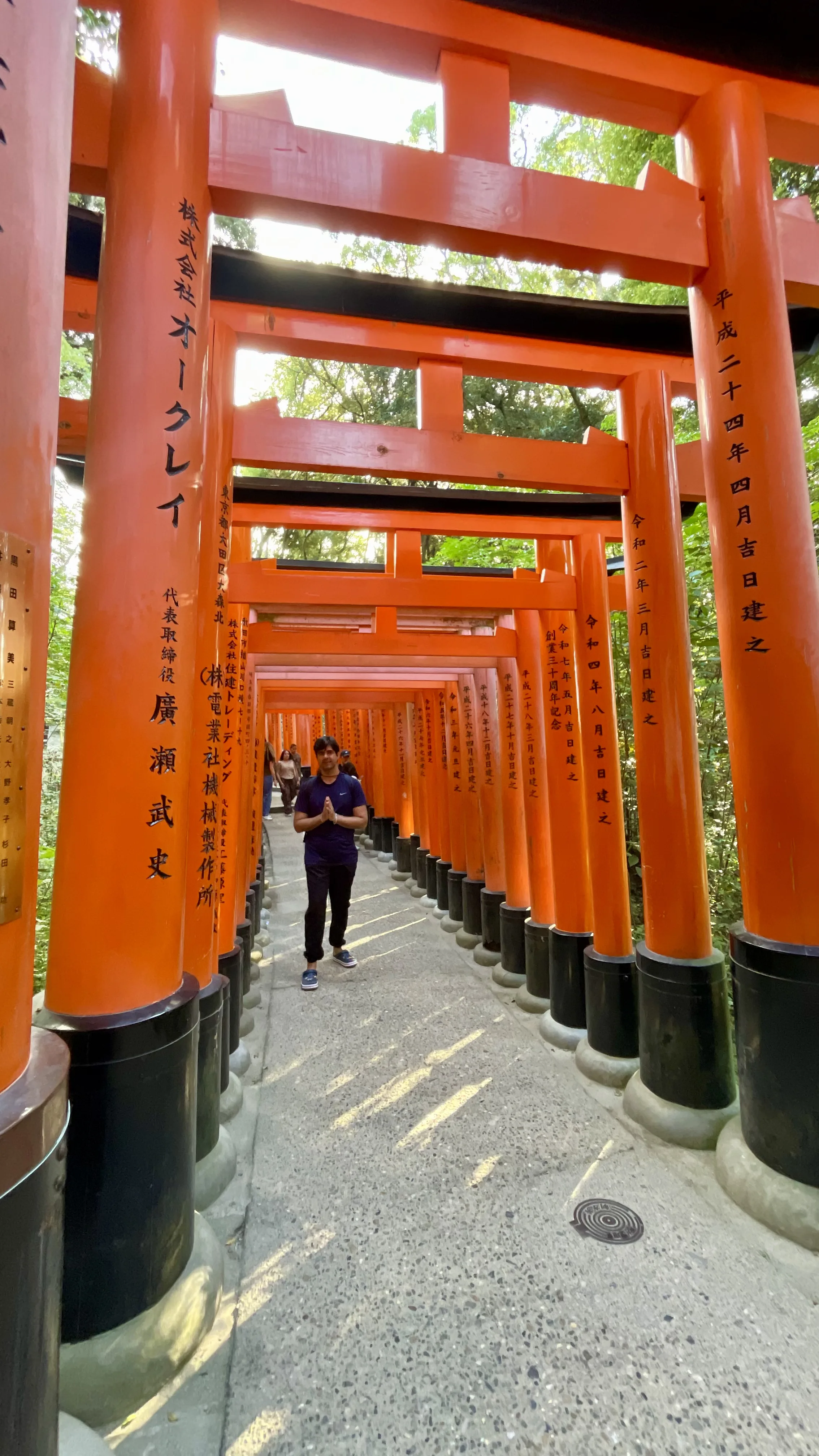 Um visitante em pé com as mãos em posição de oração no meio de um túnel alaranjado e vibrante de milhares de portões torii no Santuário Fushimi Inari, em Kyoto, com inscrições japonesas pretas em cada pilar e luz natural filtrada.