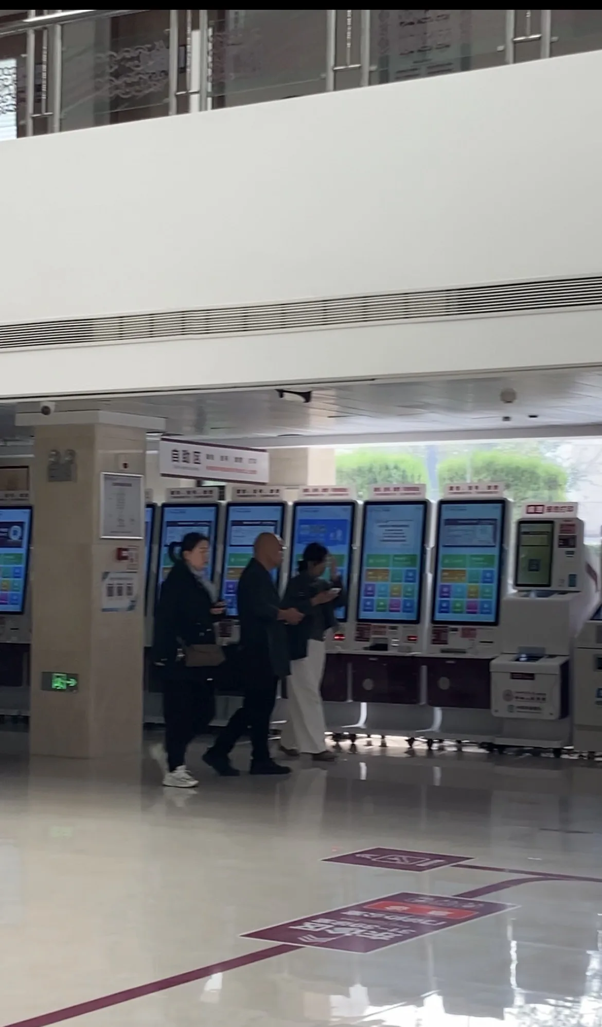Três pessoas caminhando em frente a uma fileira de totens eletrônicos de autoatendimento e pagamento no saguão de hospital na China.