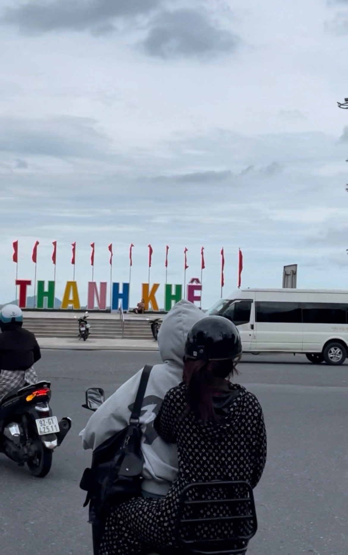 Condutores de moto em trânsito urbano com o letreiro colorido "Thanh Khê" e bandeiras ao fundo.