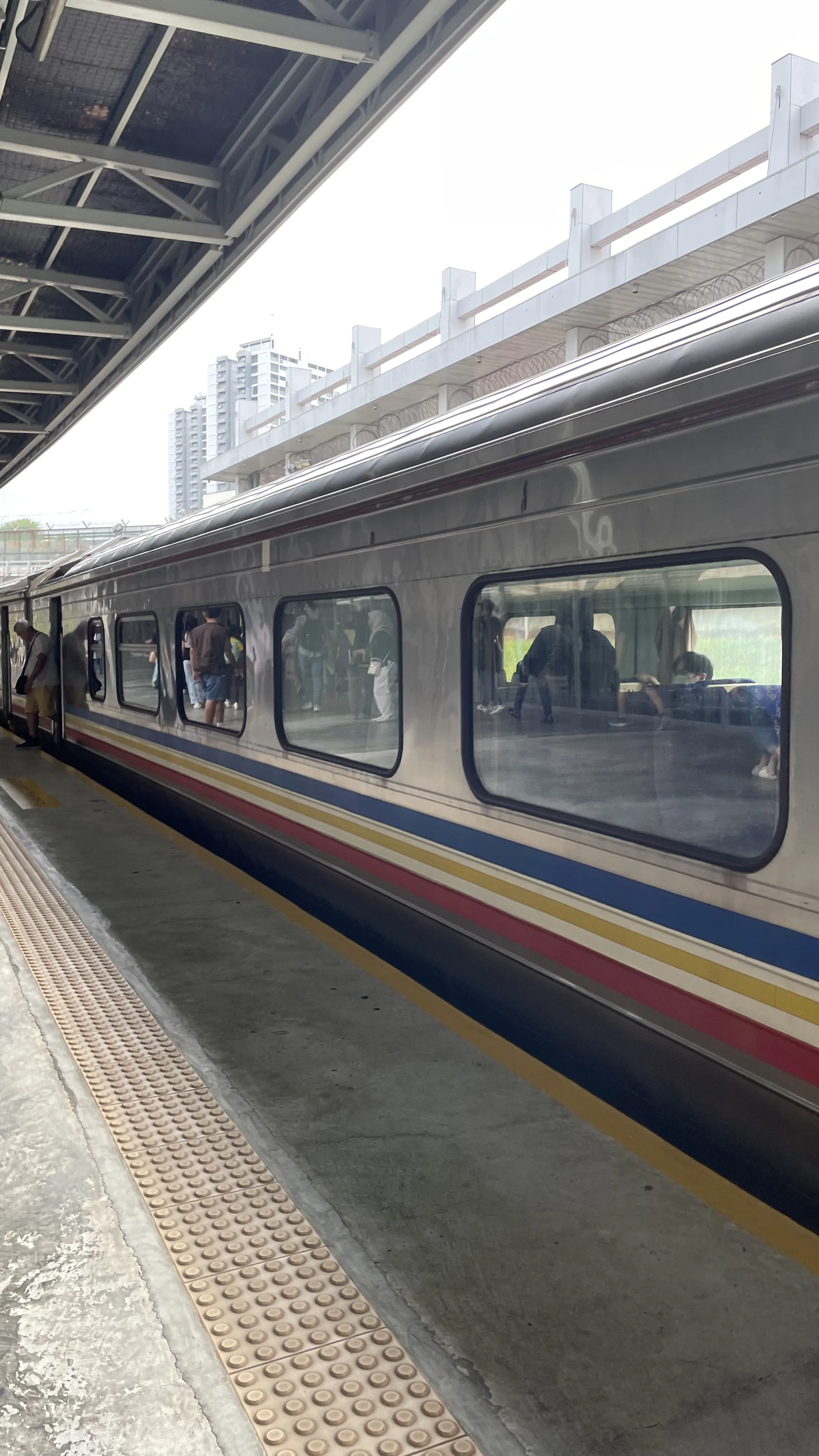 Lateral de um trem prateado com detalhes coloridos estacionado na plataforma de uma estação ferroviária.
