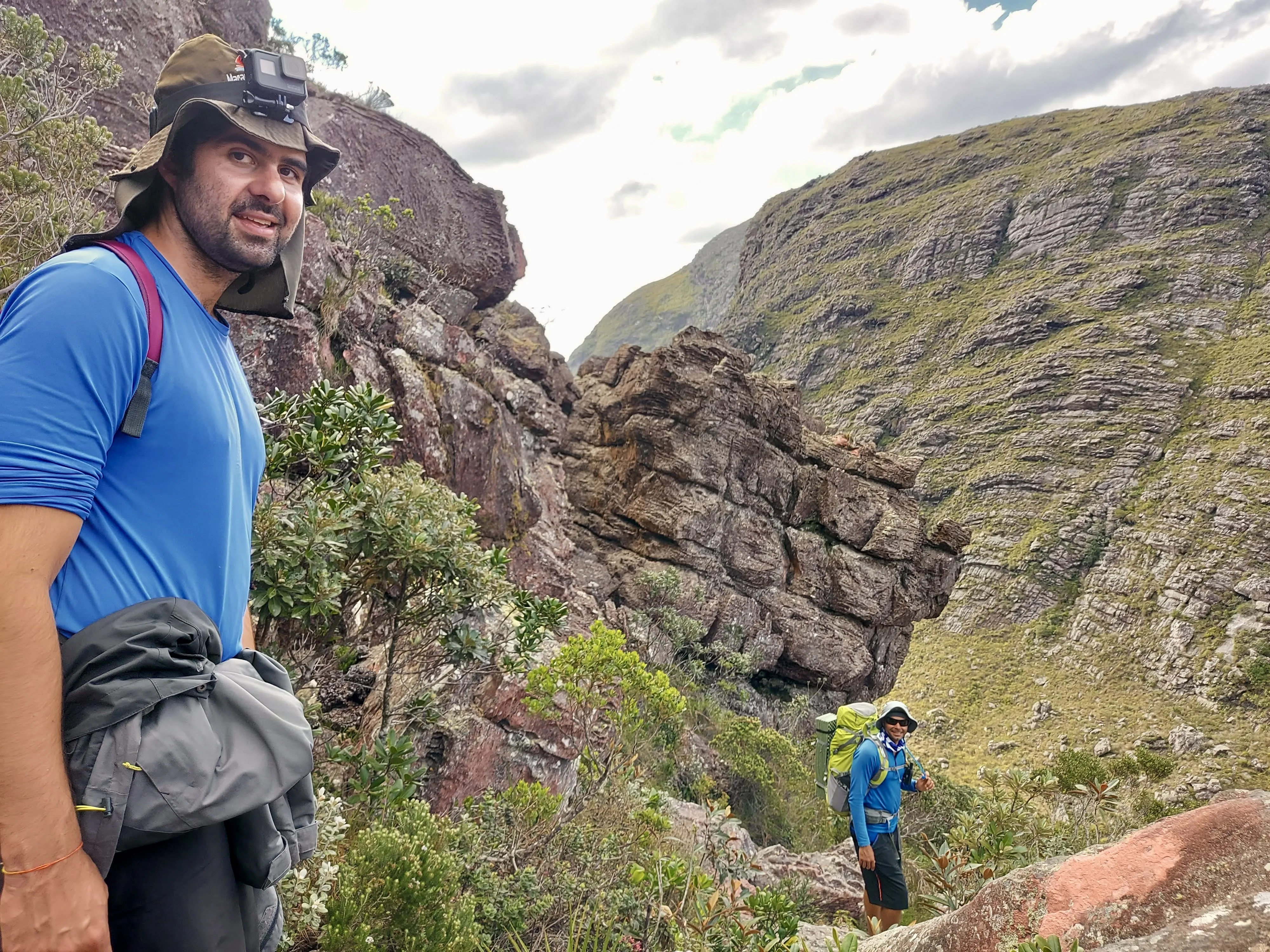 Trekking em terreno rochoso: homem sorridente com chapéu de sol e mochila em primeiro plano, olhando para a câmera. Atrás dele, em uma trilha na encosta da montanha, outro caminhante com mochila grande desce, com formações rochosas e vale verde ao fundo.