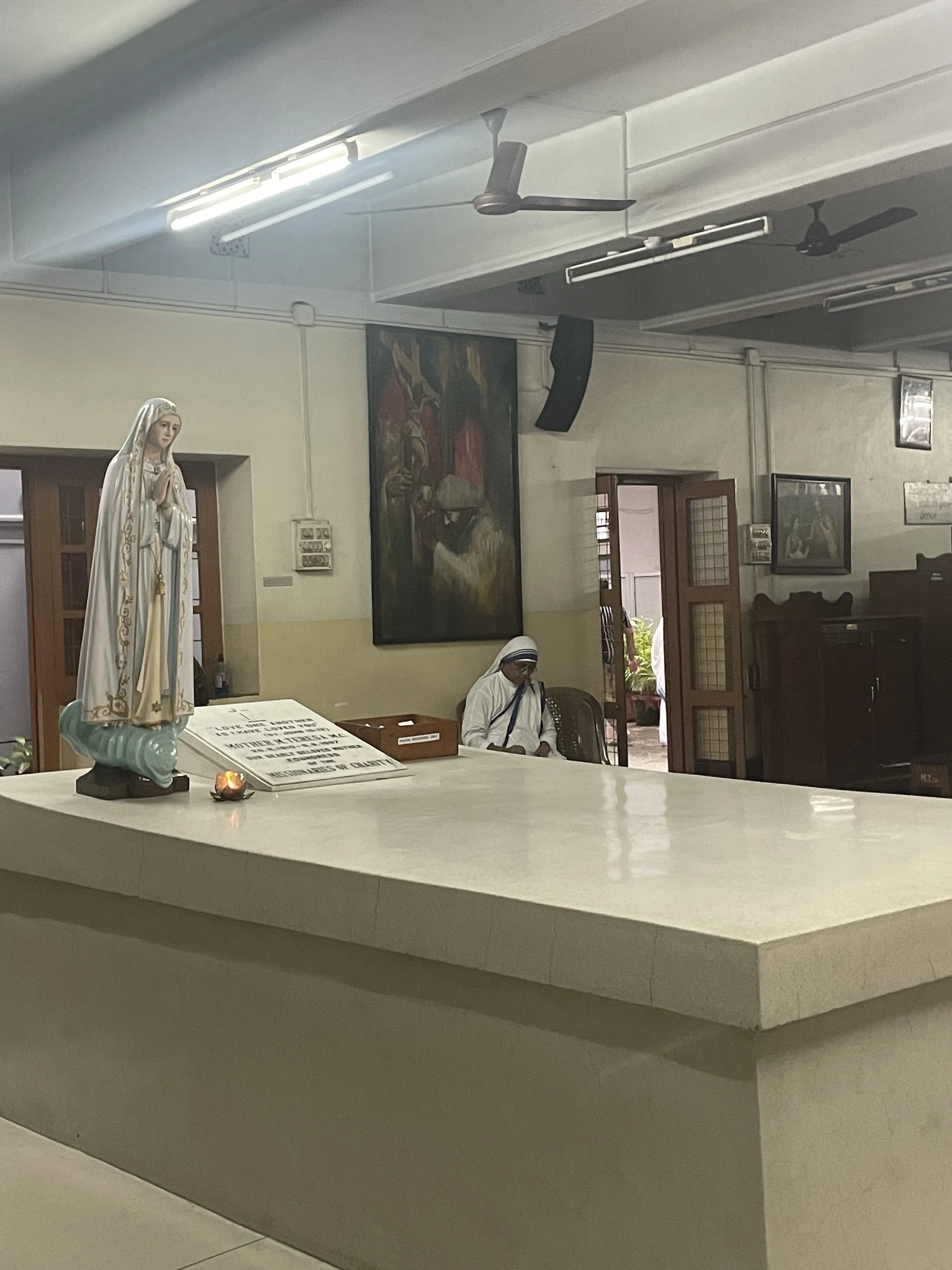 Interior tranquilo da capela na Casa Mãe das Missionárias da Caridade em Calcutá, exibindo o túmulo branco de Madre Teresa de Calcutá, uma estátua da Virgem Maria e uma freira sentada em oração ao fundo.