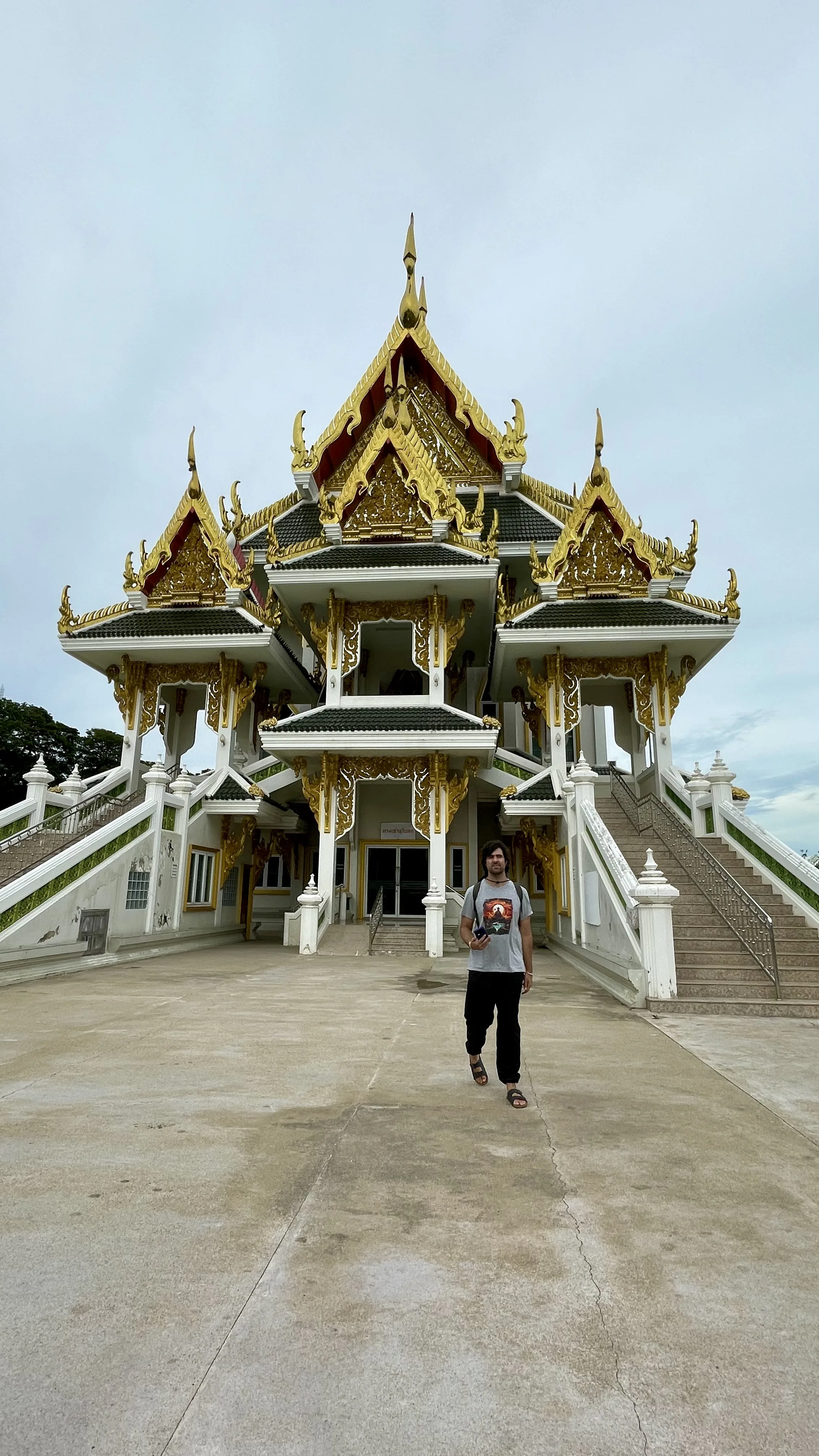Turista caminhando pela esplanada de concreto em direção à fachada frontal do moderno e suntuoso Ubosot de Wat Khun Inthapramun, destacando as escadarias laterais e a rica decoração dourada dos múltiplos telhados.