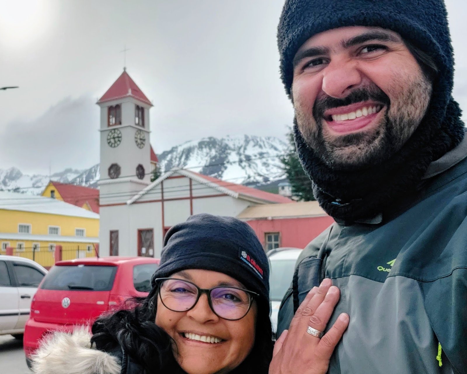 Um homem e uma mulher sorridentes em selfie na rua, tendo ao fundo uma igreja com torre vermelha e montanhas nevadas em Ushuaia.