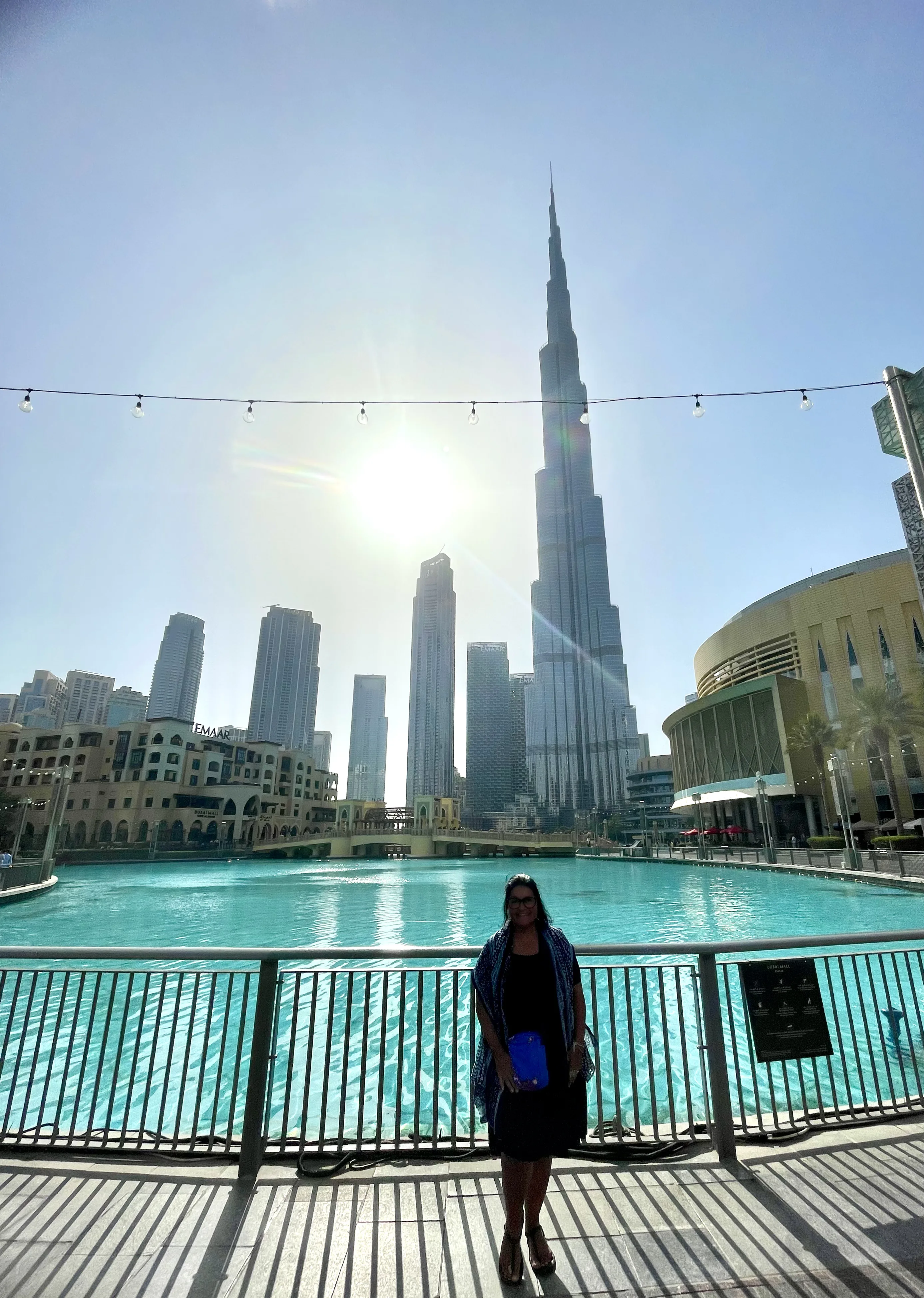 Mulher de vestido, em pé posando sorridente na esplanada à beira da grande piscina refletora, tendo ao fundo a imponente torre Burj Khalifa e os arranha-céus modernos de Dubai sob o sol forte.