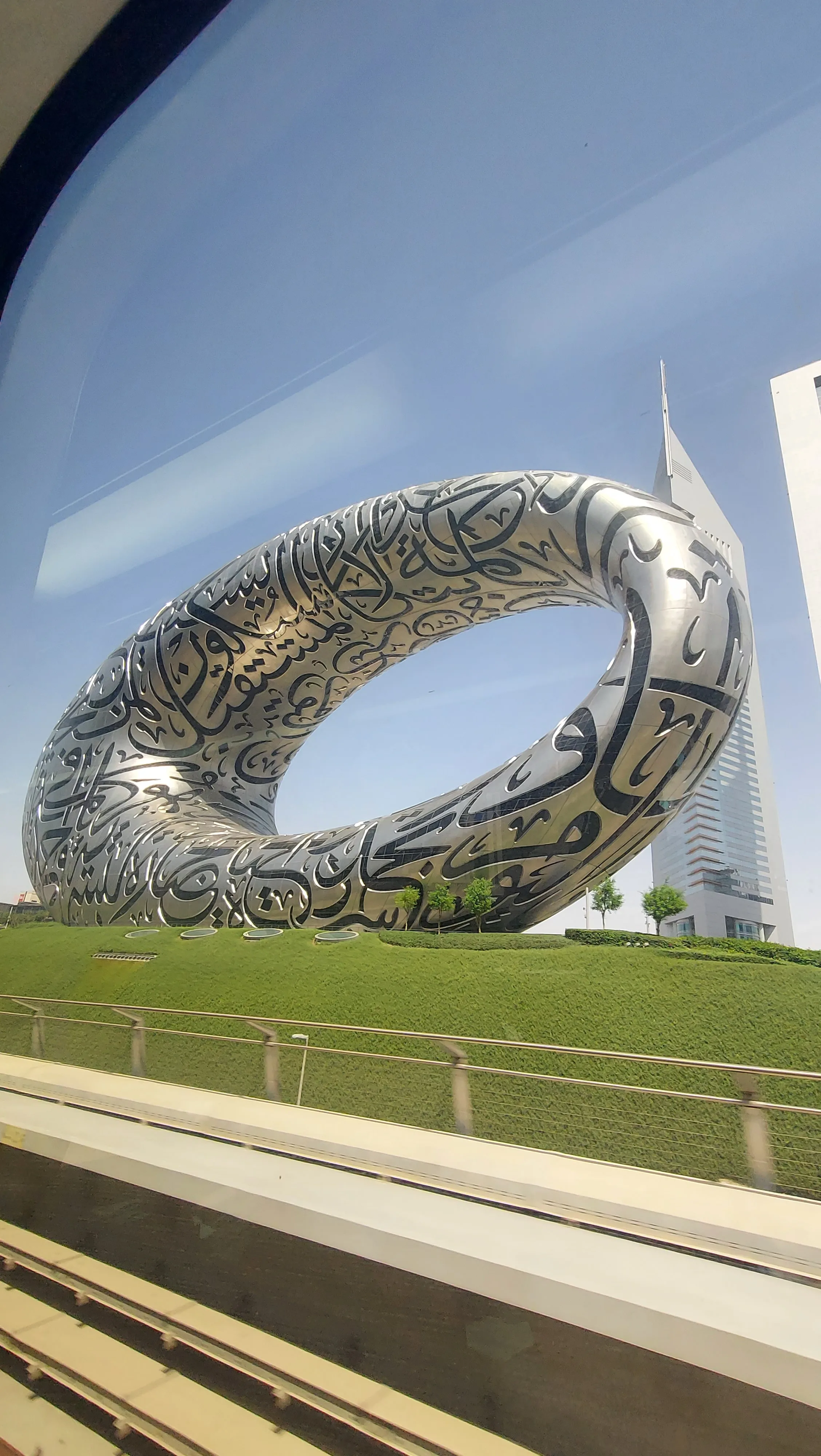 Vista externa do icônico edifício futurista do Museu do Futuro em Dubai, com sua chamativa estrutura em formato de toro oval revestida de aço inoxidável e inscrições em caligrafia árabe, erguendo-se sobre uma colina gramada.