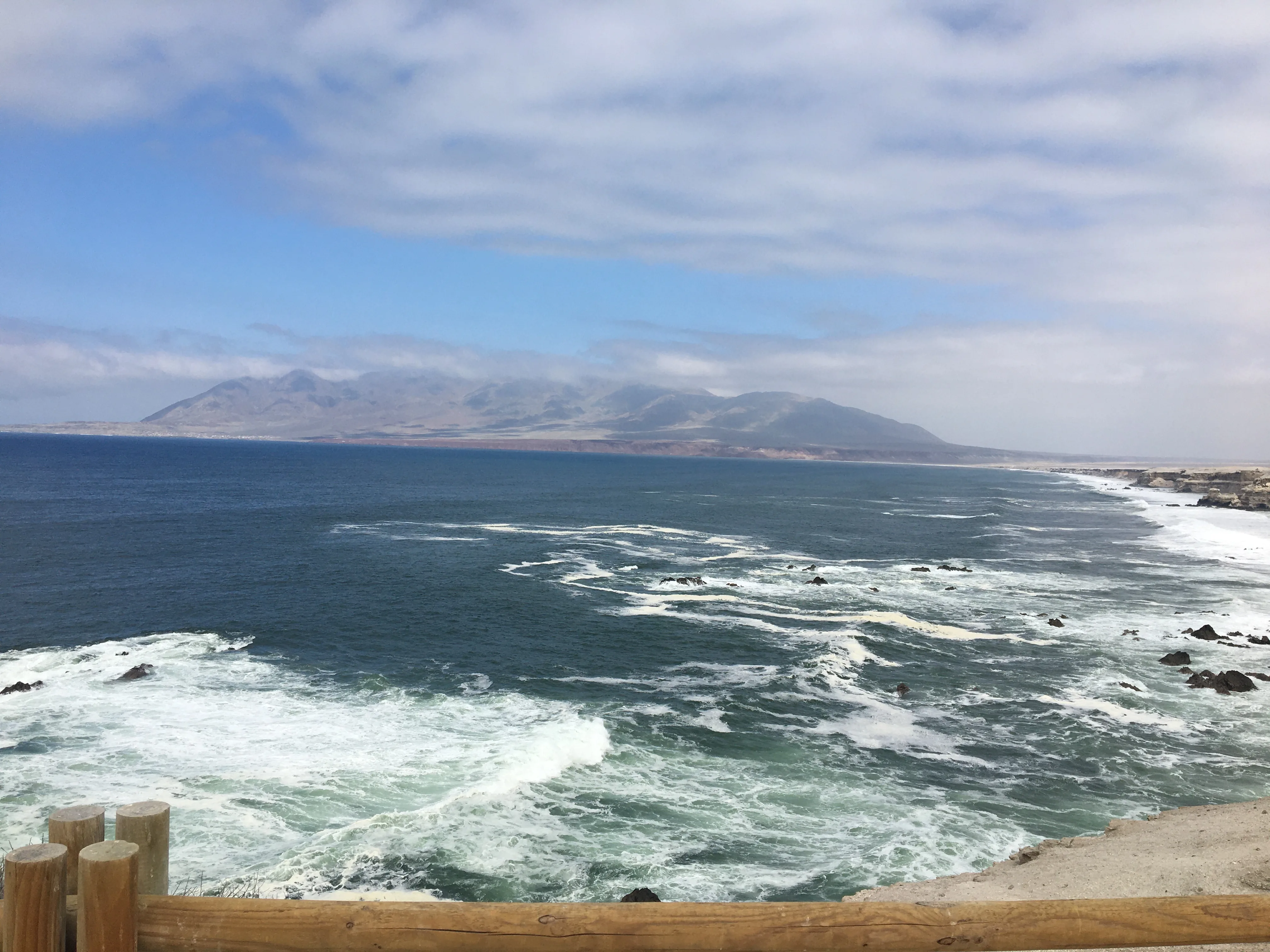 Vista panorâmica do Oceano Pacífico a partir da costa de Antofagasta, com ondas espumosas quebrando nas rochas e montanhas áridas ao fundo sob um céu nublado.