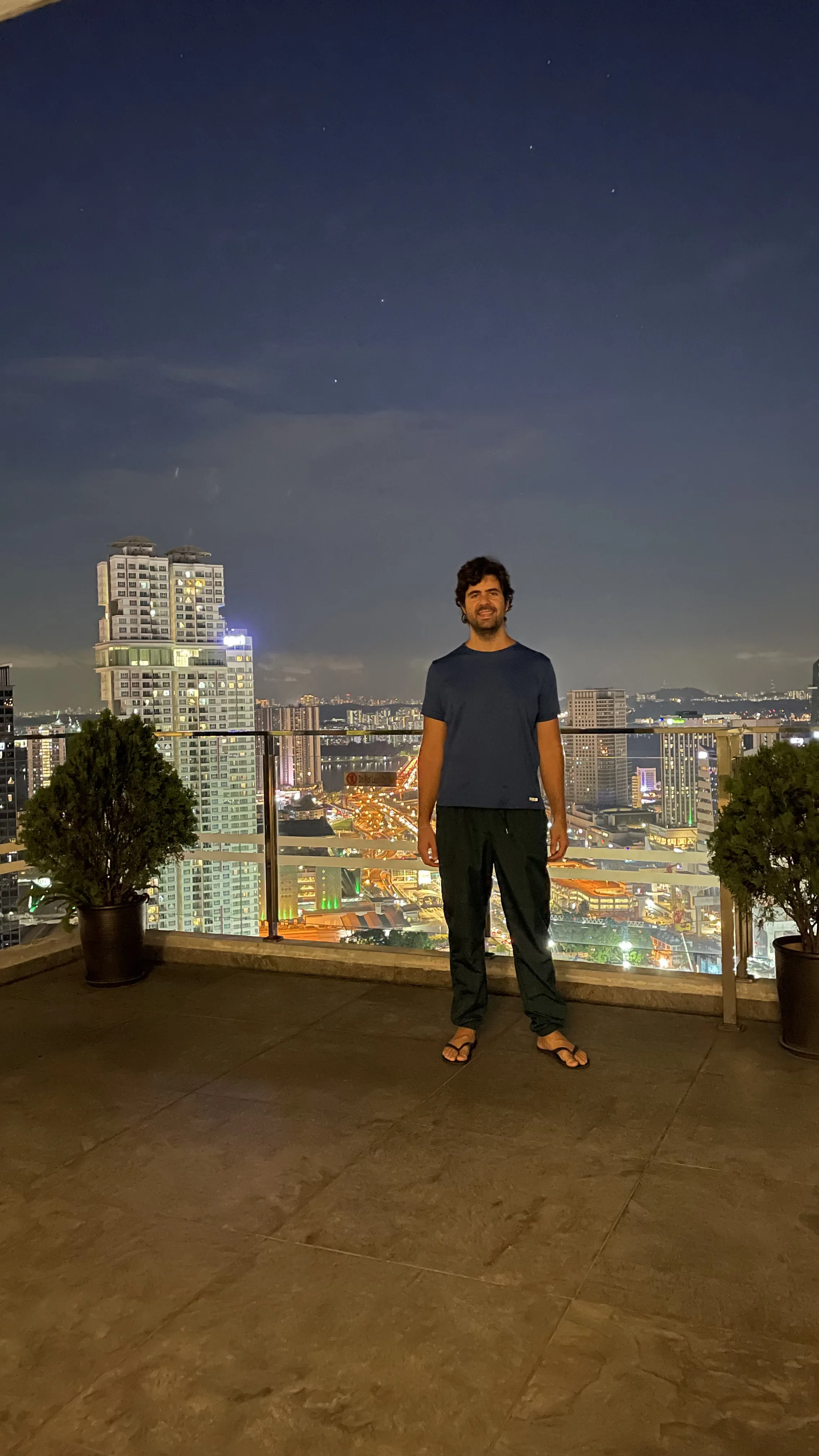 Um homem em pé no terraço de um edifício, posando com a paisagem urbana iluminada de Johor Bahru à noite ao fundo.