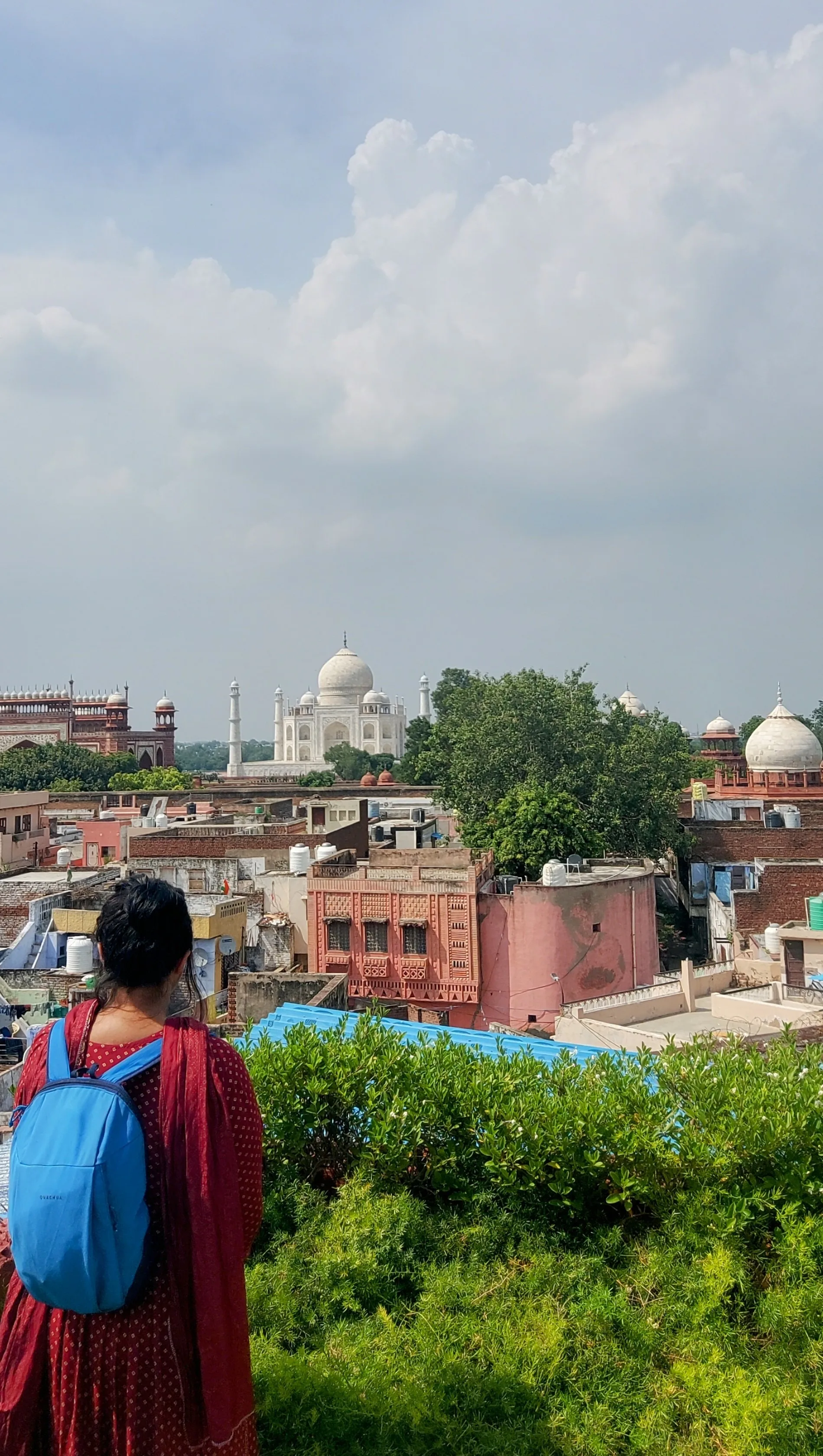 Visitante com mochila azul e blusa vermelha contemplando a vista panorâmica do Taj Mahal e dos telhados da cidade a partir do terraço de um restaurante local cercado por vegetação.