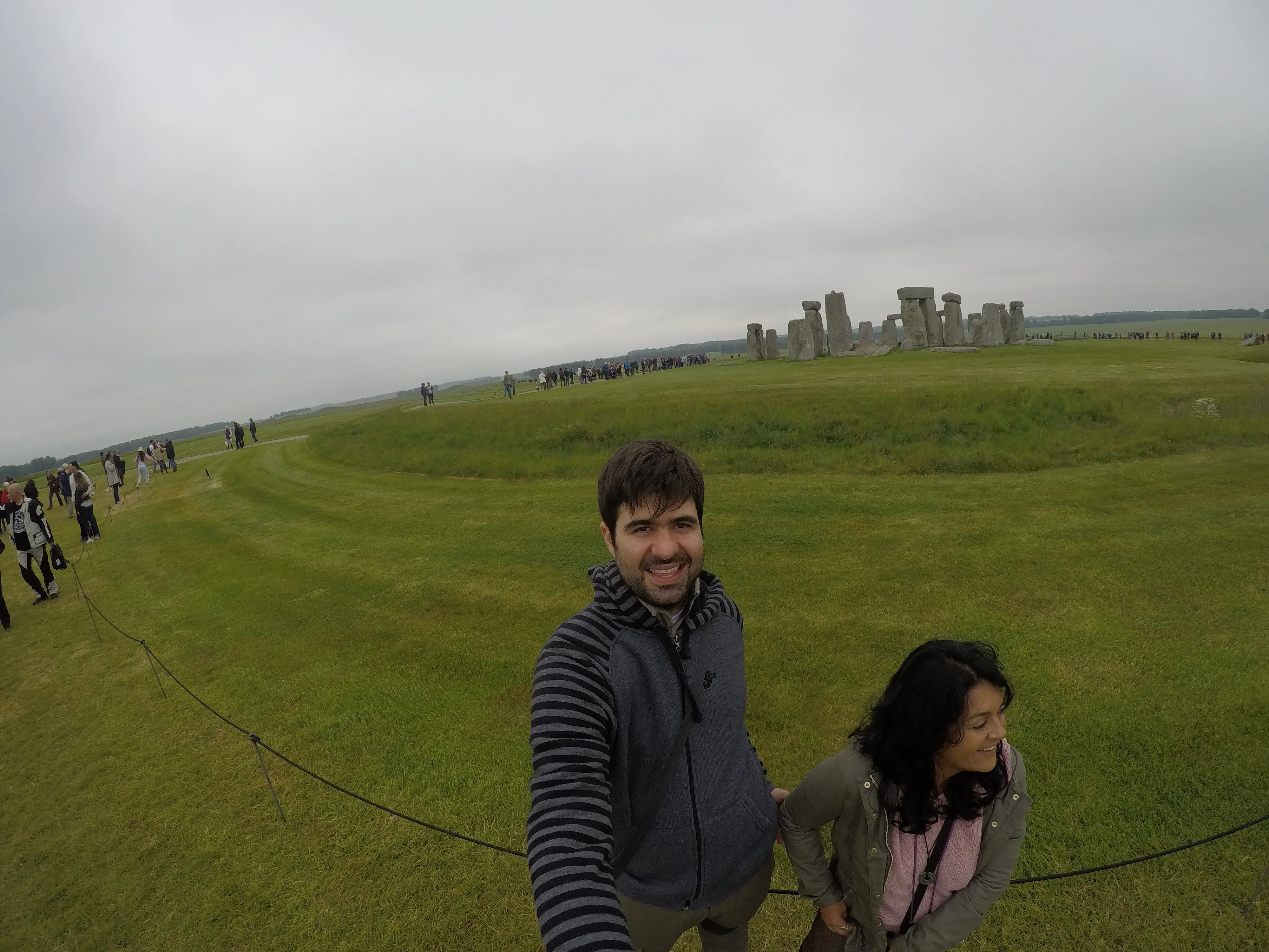 Casal de viajantes sorrindo no gramado com o círculo de pedras de Stonehenge ao longe na Inglaterra.