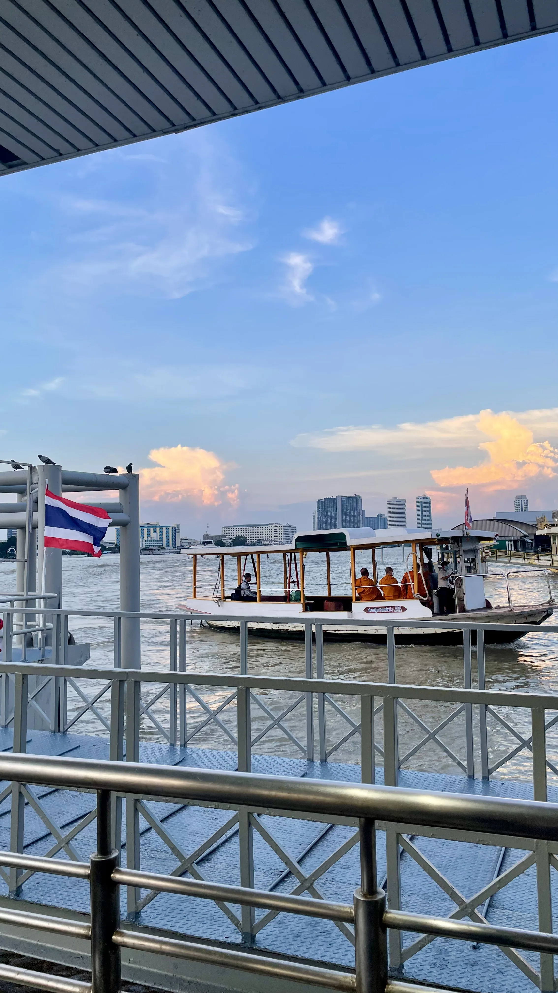Barco de passageiros navegando pelo rio Chao Phraya em Banguecoque, visto a partir de uma plataforma de embarque com a bandeira da Tailândia.