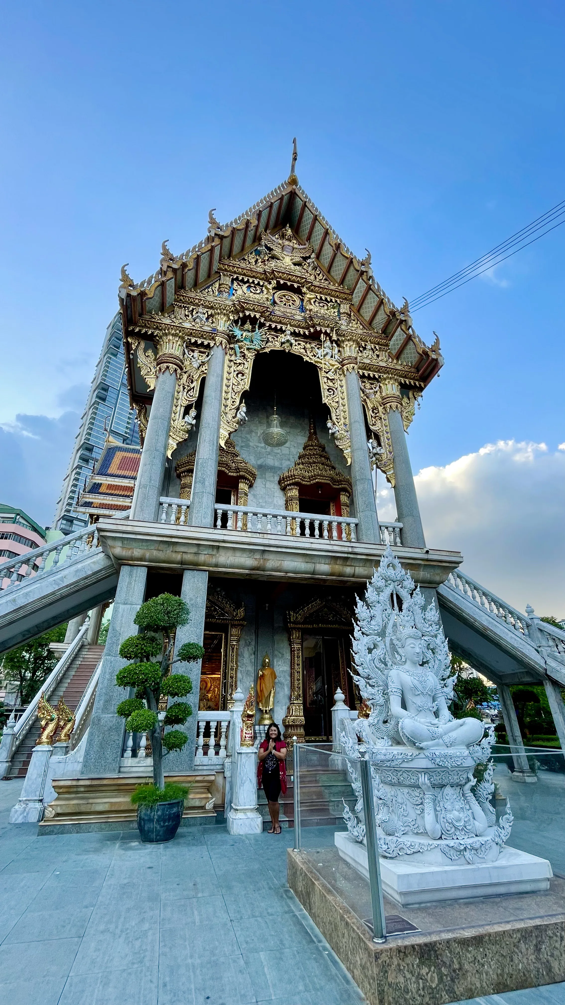 Uma mulher em pé com as mãos juntas em reverência diante da arquitetura tailandesa e estátua de divindade no Wat Suthiwaram