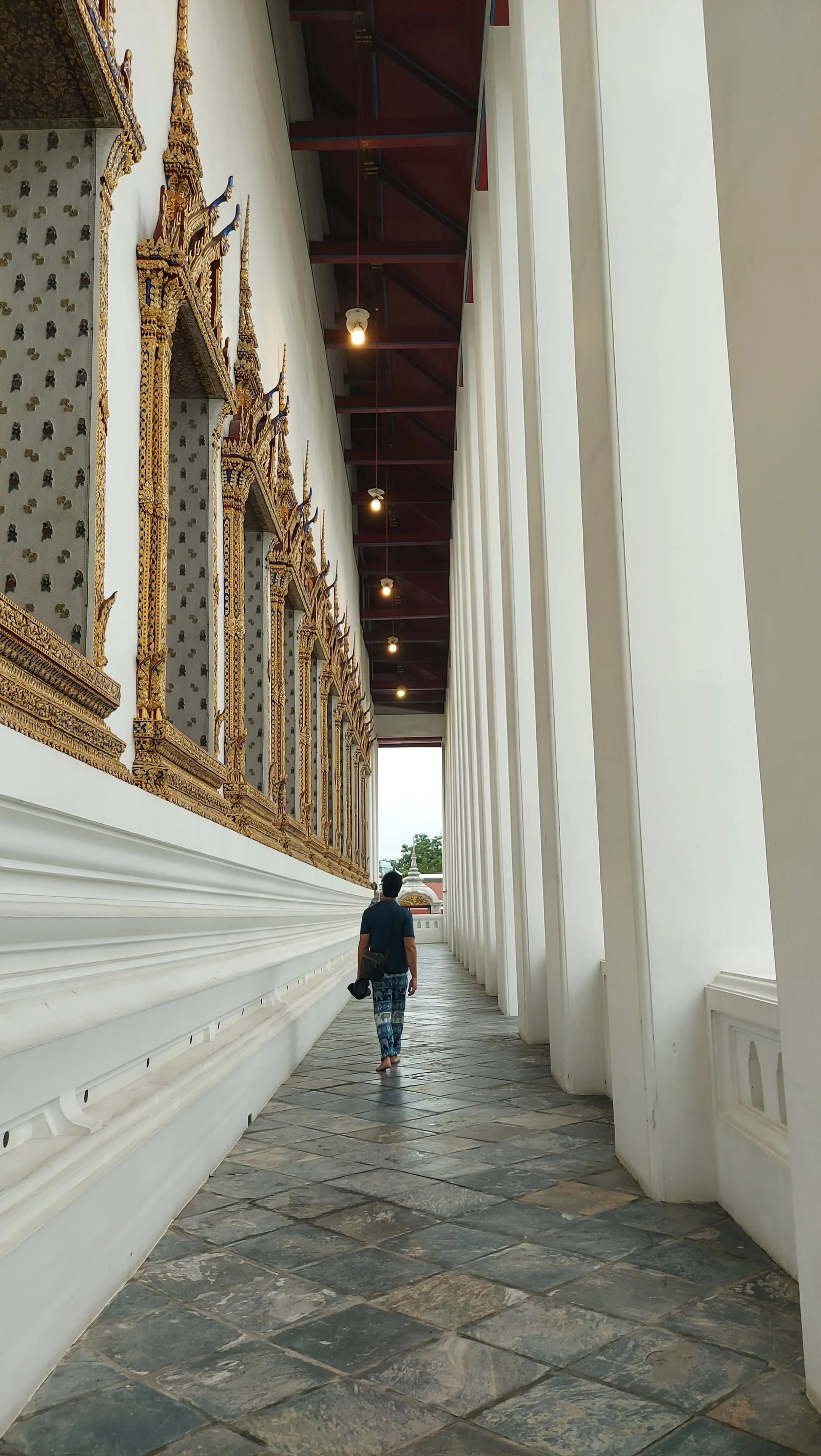Um homem caminhando ao longo do corredor com colunatas brancas e paredes decoradas do Wat Suthat em Banguecoque.