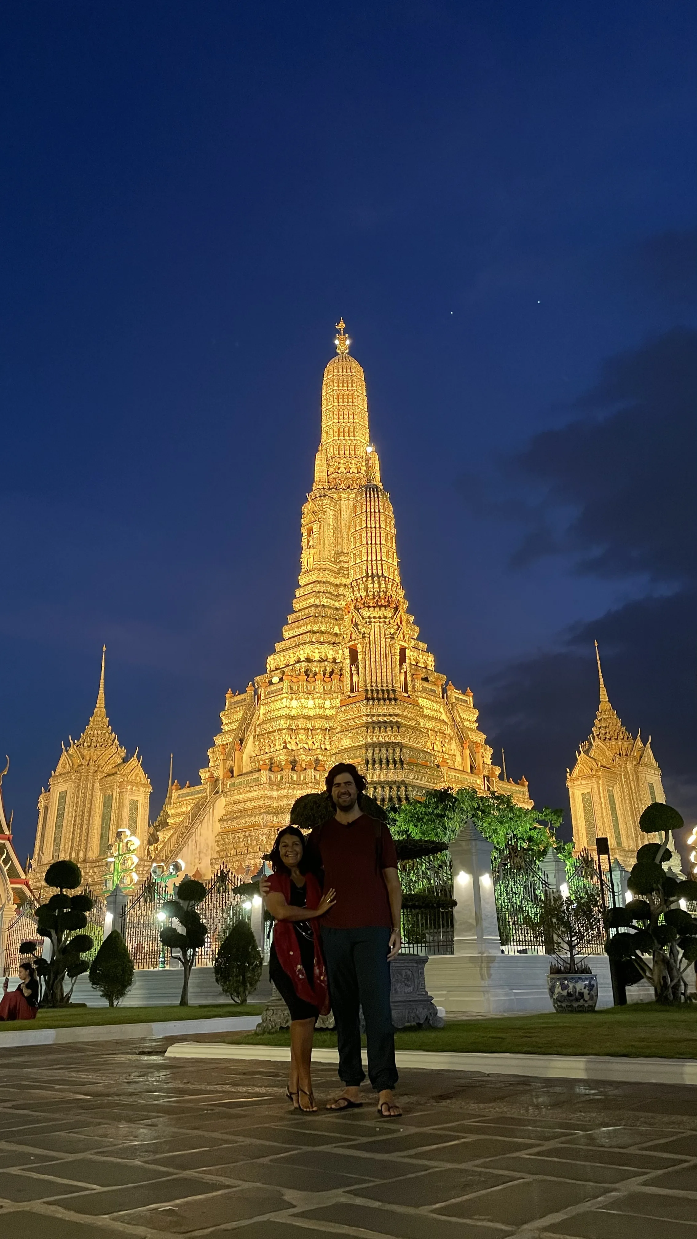 Casal sorridente em pé em frente ao Wat Arun iluminado à noite em Banguecoque.