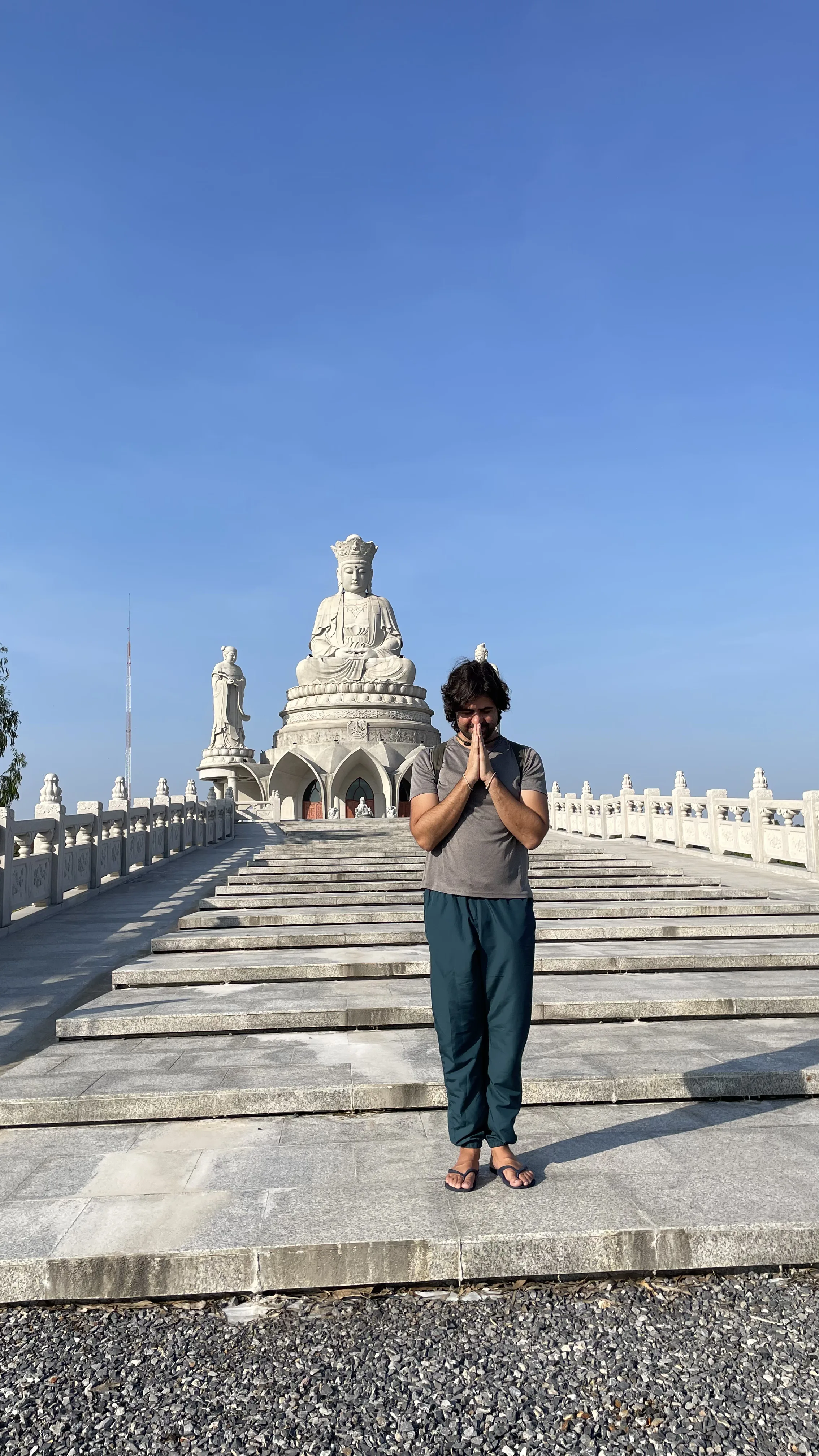Homem em pé com as mãos juntas em gesto de reverência na escadaria do templo Wihan Sadet Pho Kuanin, na Tailândia, tendo a estátua branca ao fundo.